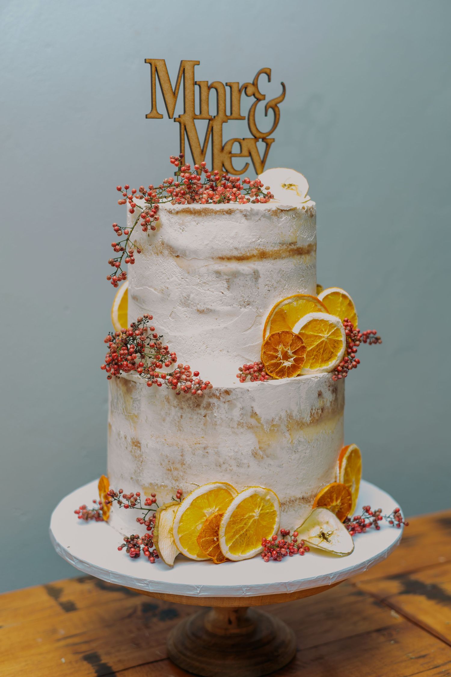 Two-tier naked wedding cake decorated with dried orange slices, pink berries, and a gold Mr and Mrs cake topper.
