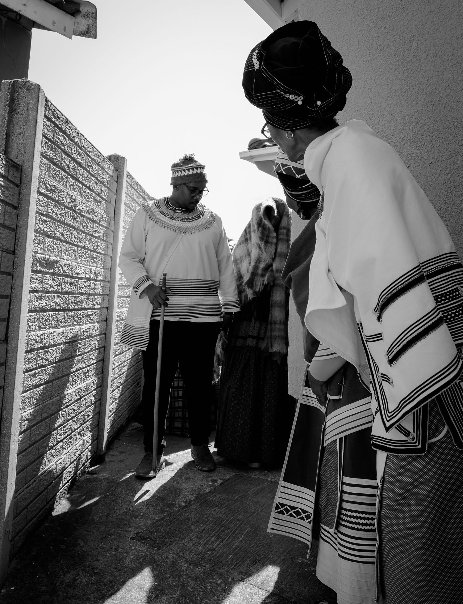 Black and white photo of two people in traditional Xhosa attire walking through a narrow outdoor passage.