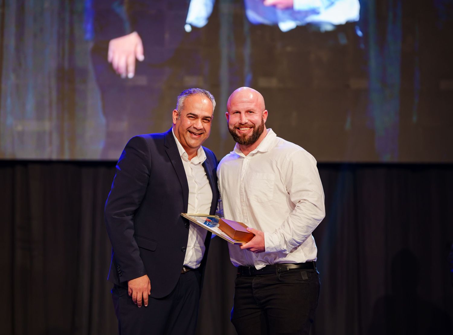 Two men share a handshake while presenting an award on stage at corporate ceremony.