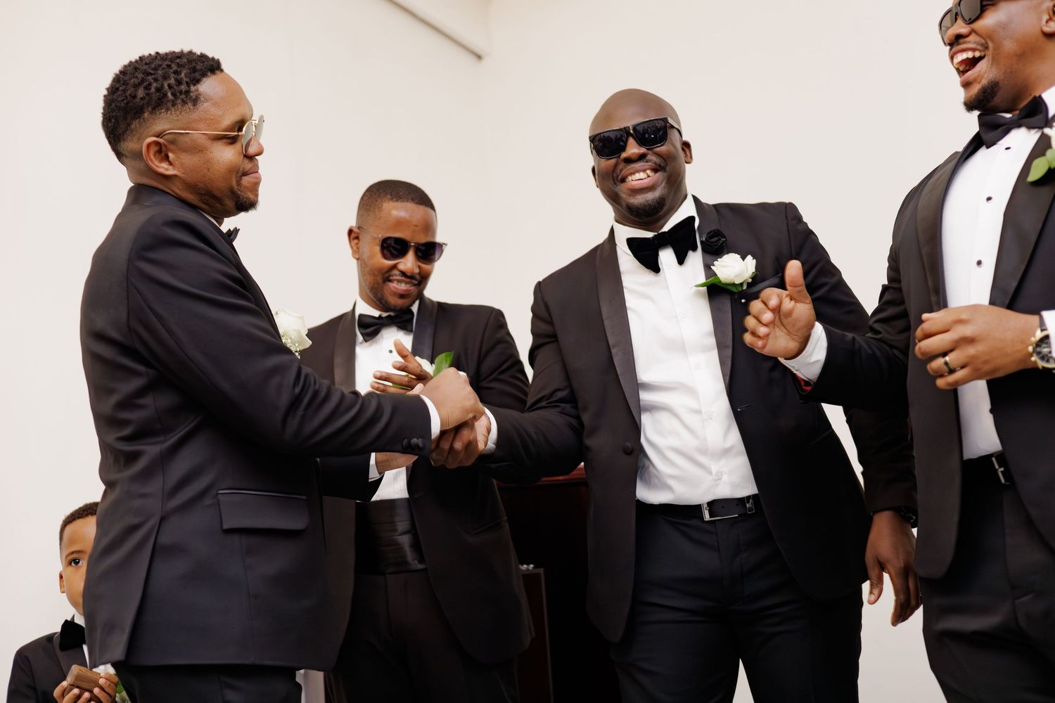 Groomsmen in black tuxedos laughing and shaking hands before wedding ceremony.