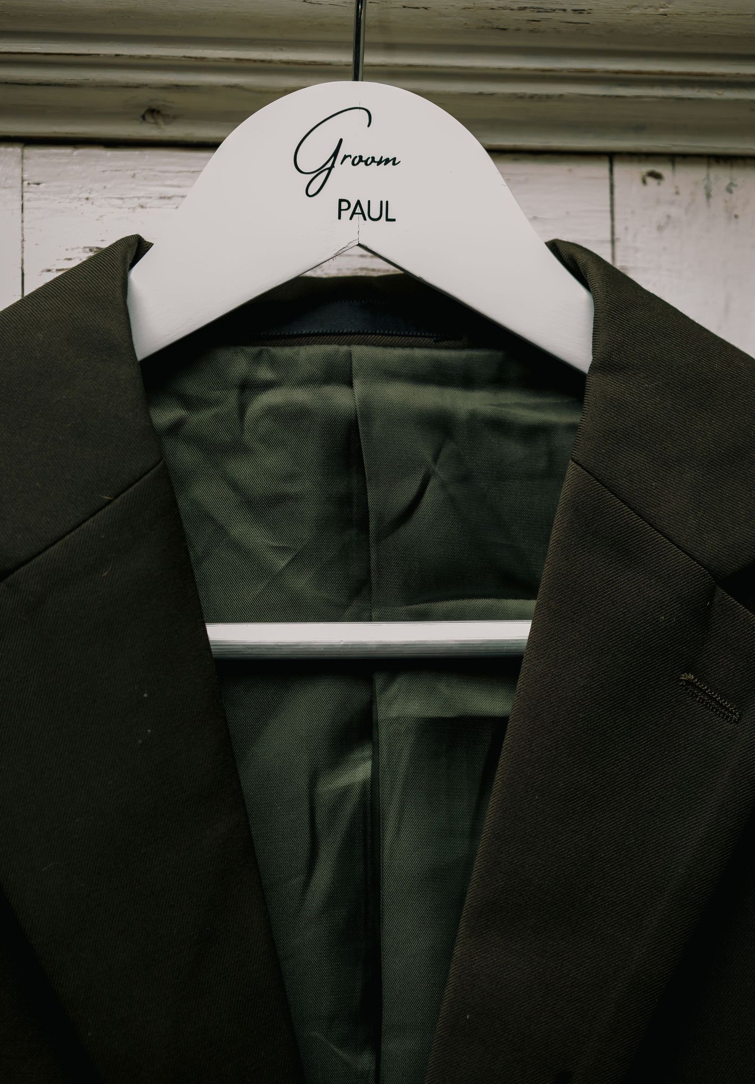 Dark green groom suit on personalized white hanger labeled Paul for wedding day.