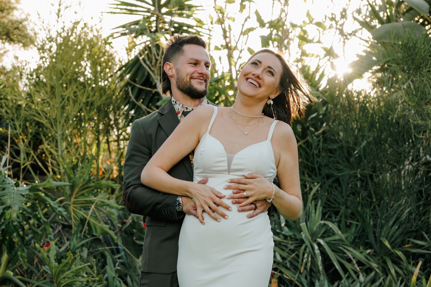 Groom embracing smiling bride in white dress surrounded by lush green foliage.