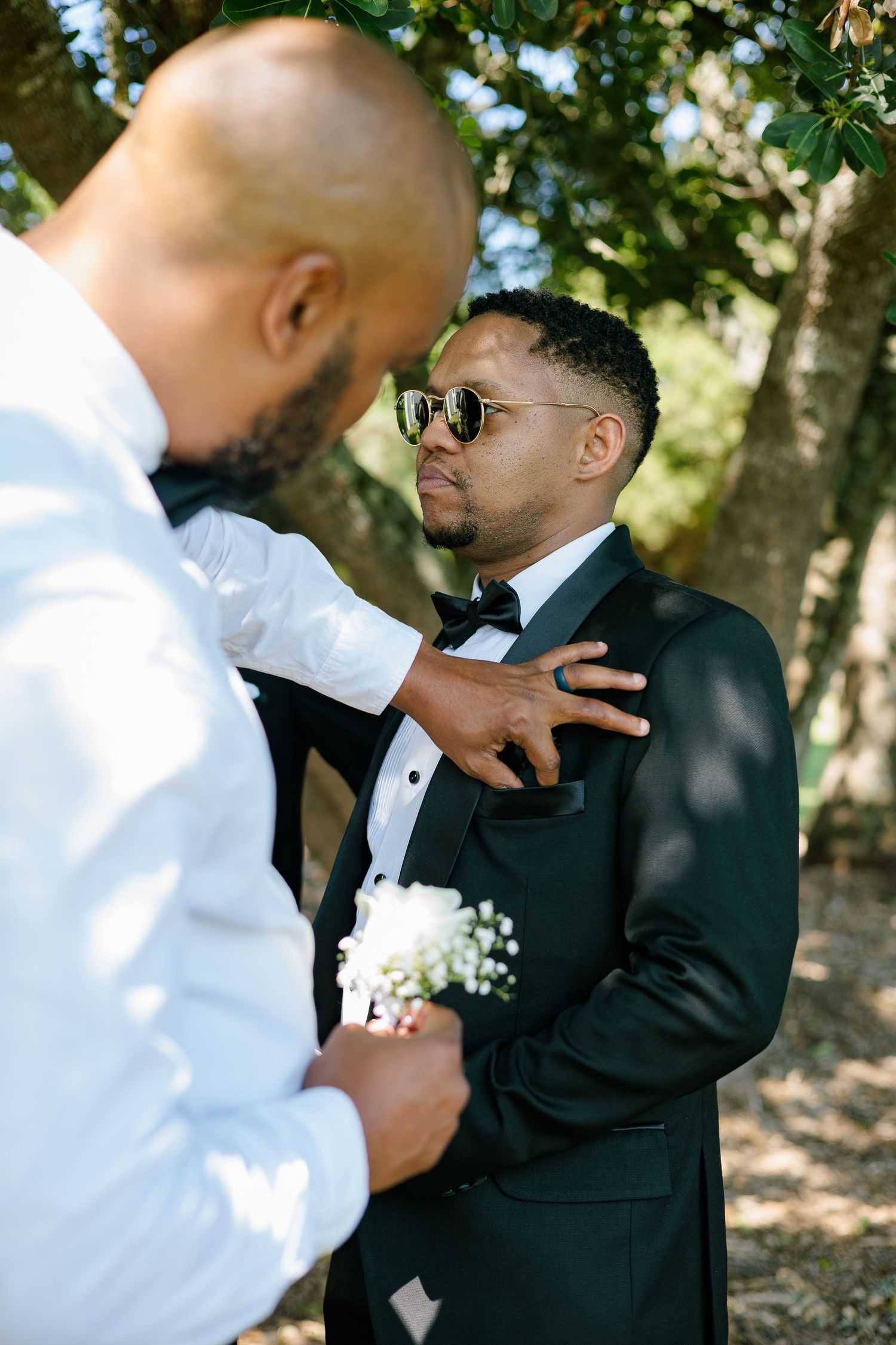 Groomsman adjusting boutonniere on groom in black tuxedo with bow tie outdoors under trees.