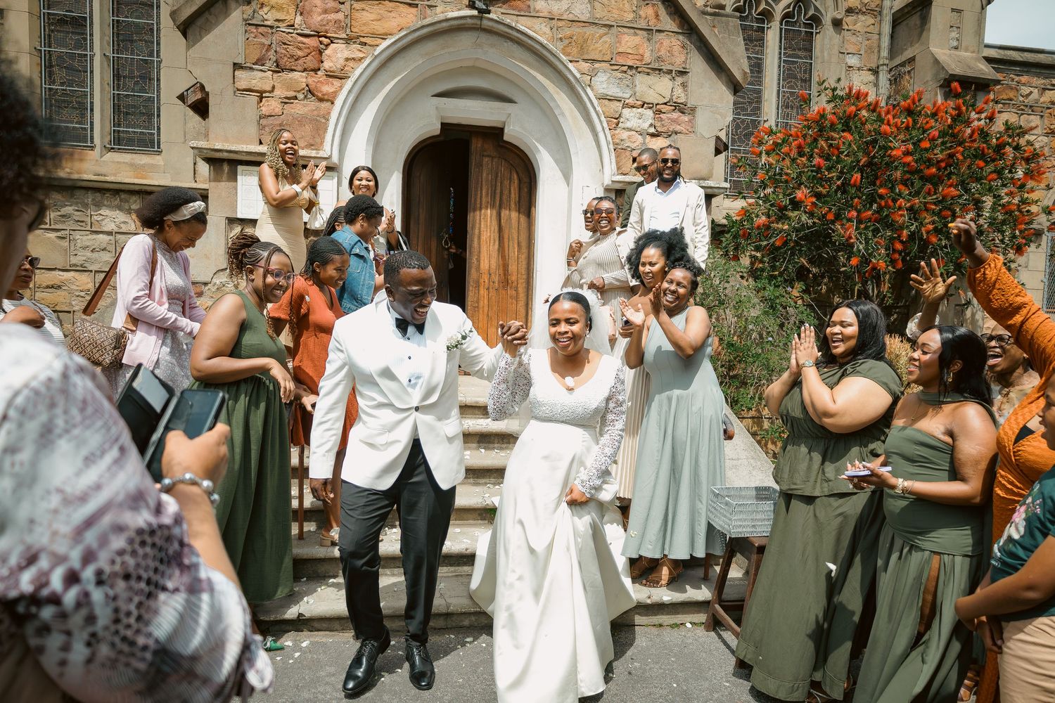 Happy newlyweds walk hand in hand outside stone church with celebrating wedding party.