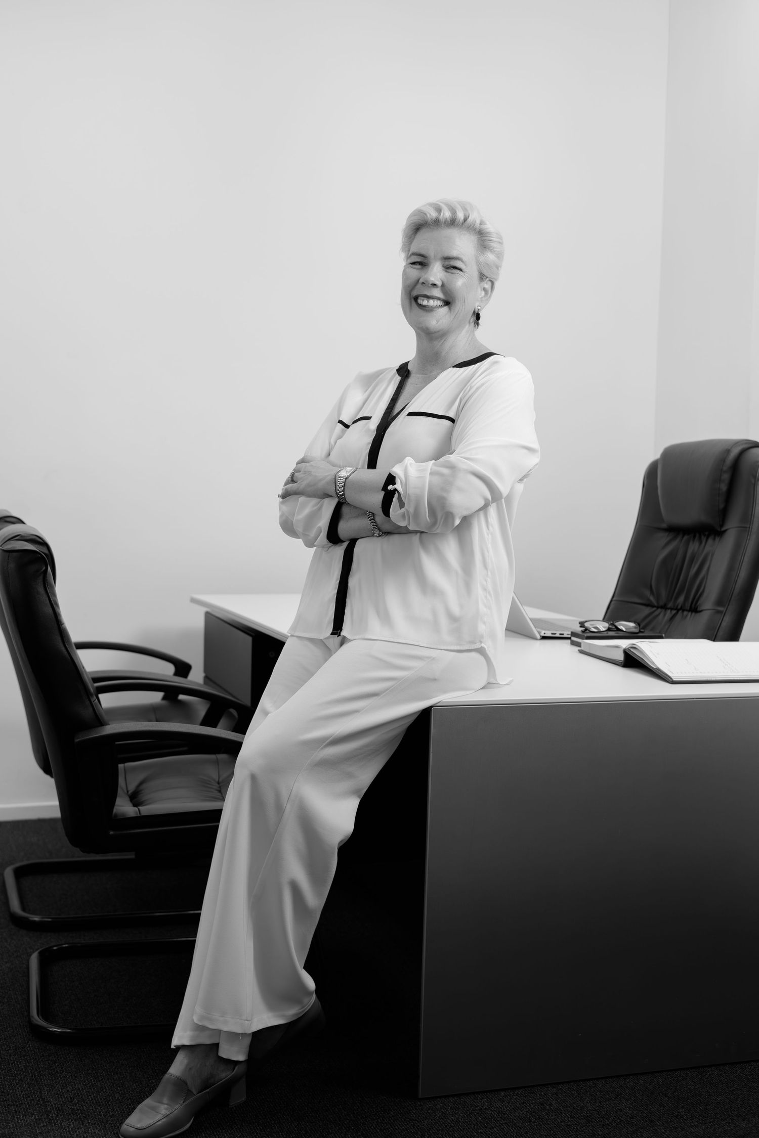 Confident businesswoman in white suit leans against office desk smiling.