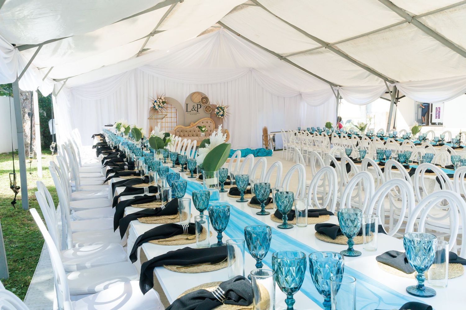 Luxurious wedding reception tent with long tables set in teal and gold, featuring blue goblets and white chairs.
