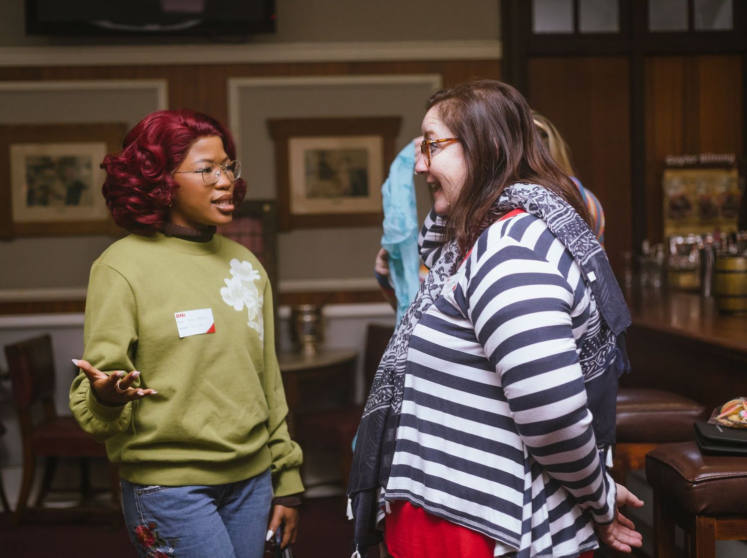 Two women smiling and conversing at an indoor networking event, one in green sweatshirt, one in stripes.