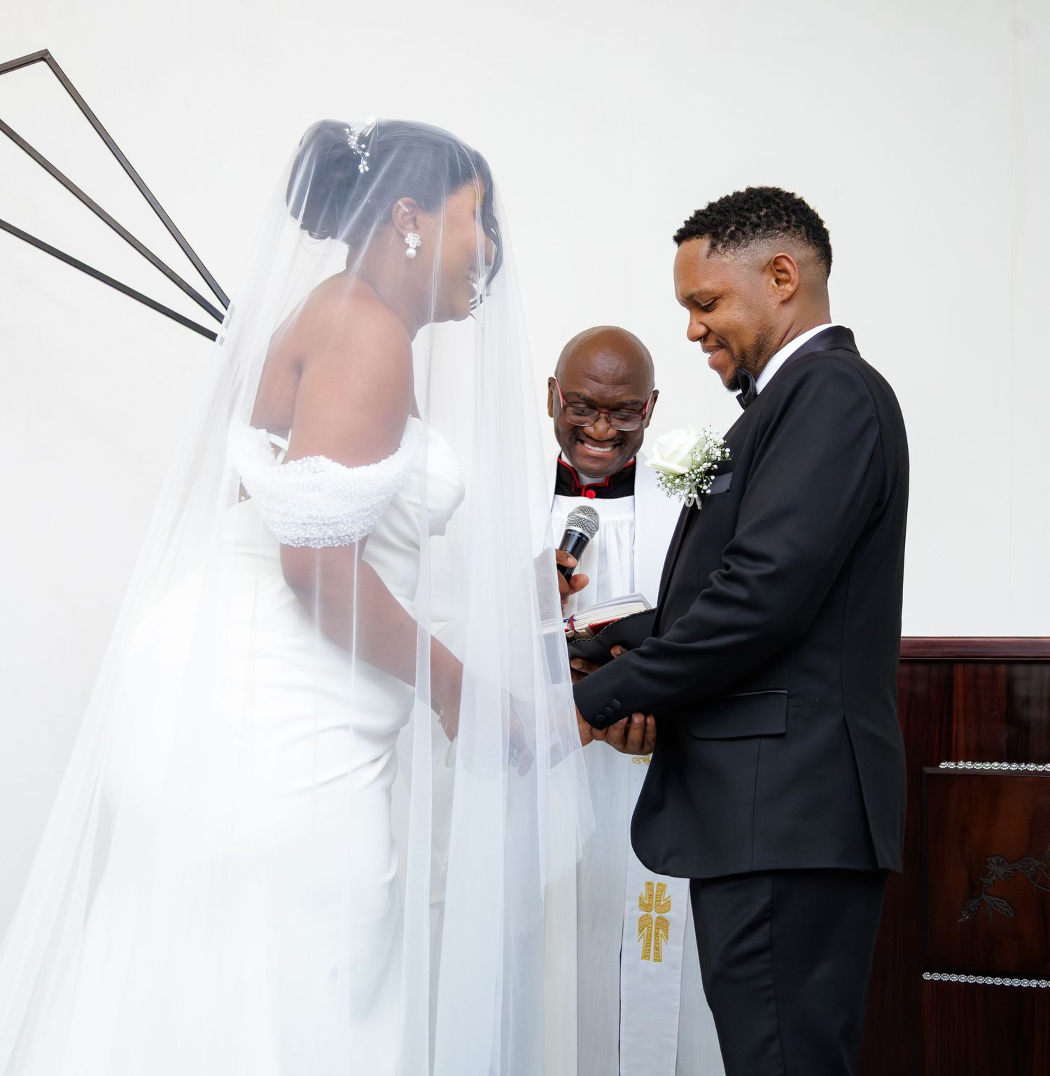 Groom holds bride's hands during wedding vows as smiling officiant reads ceremony in elegant church.