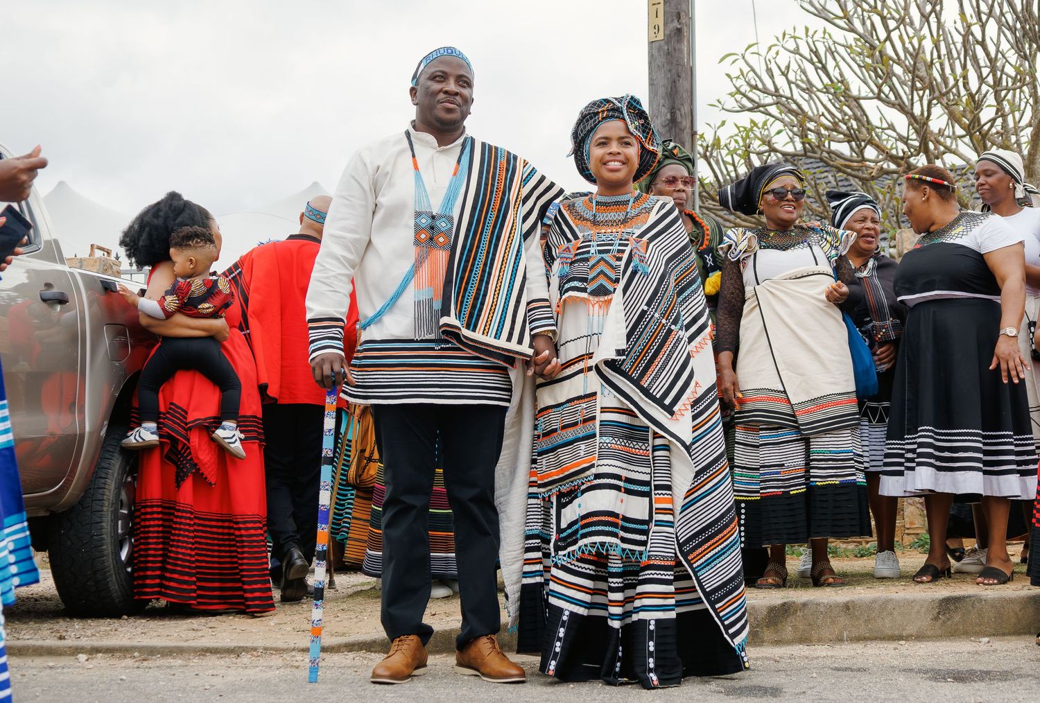 A couple stands proudly in traditional Xhosa striped ceremonial attire at an outdoor cultural gathering.