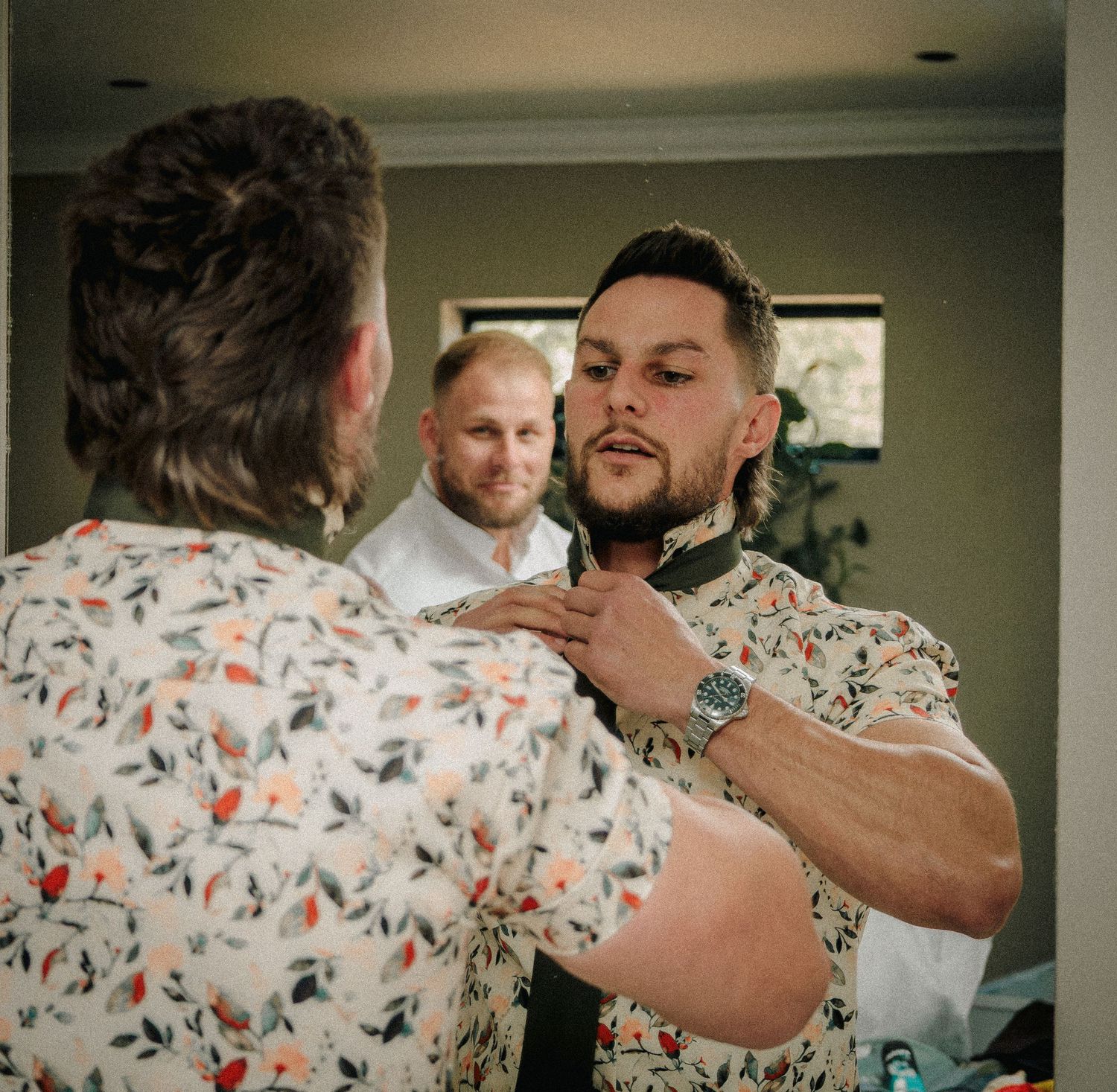 Groomsmen helping groom adjust his tie before wedding ceremony in floral shirts.