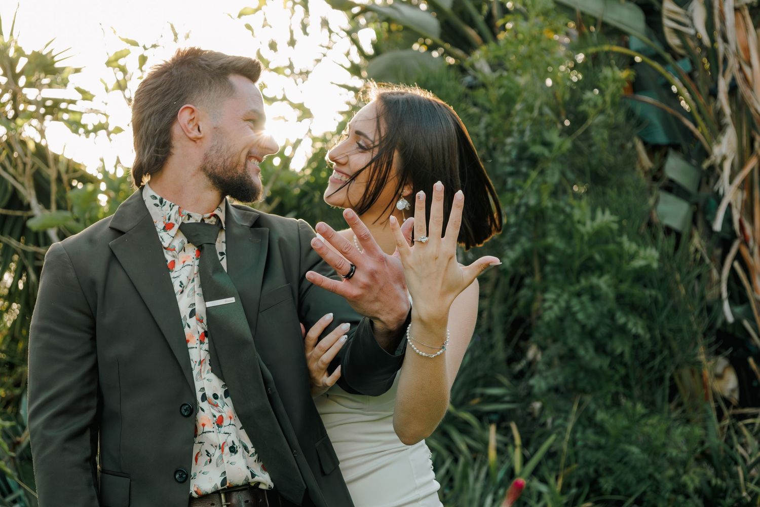 Happy couple showing off wedding rings while laughing in tropical garden setting.