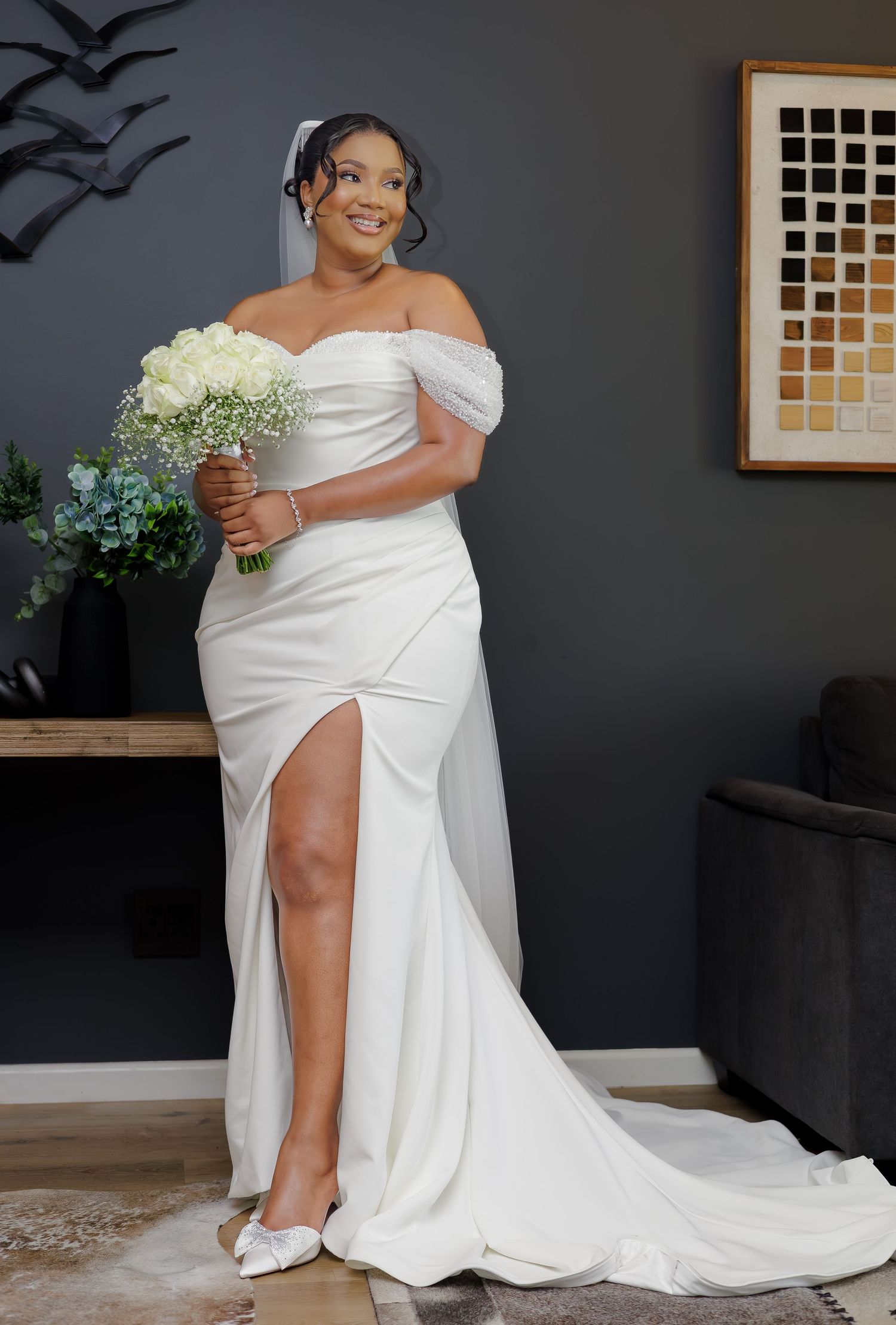 Beautiful bride in fitted off-shoulder white gown with slit holding white bouquet before wedding.