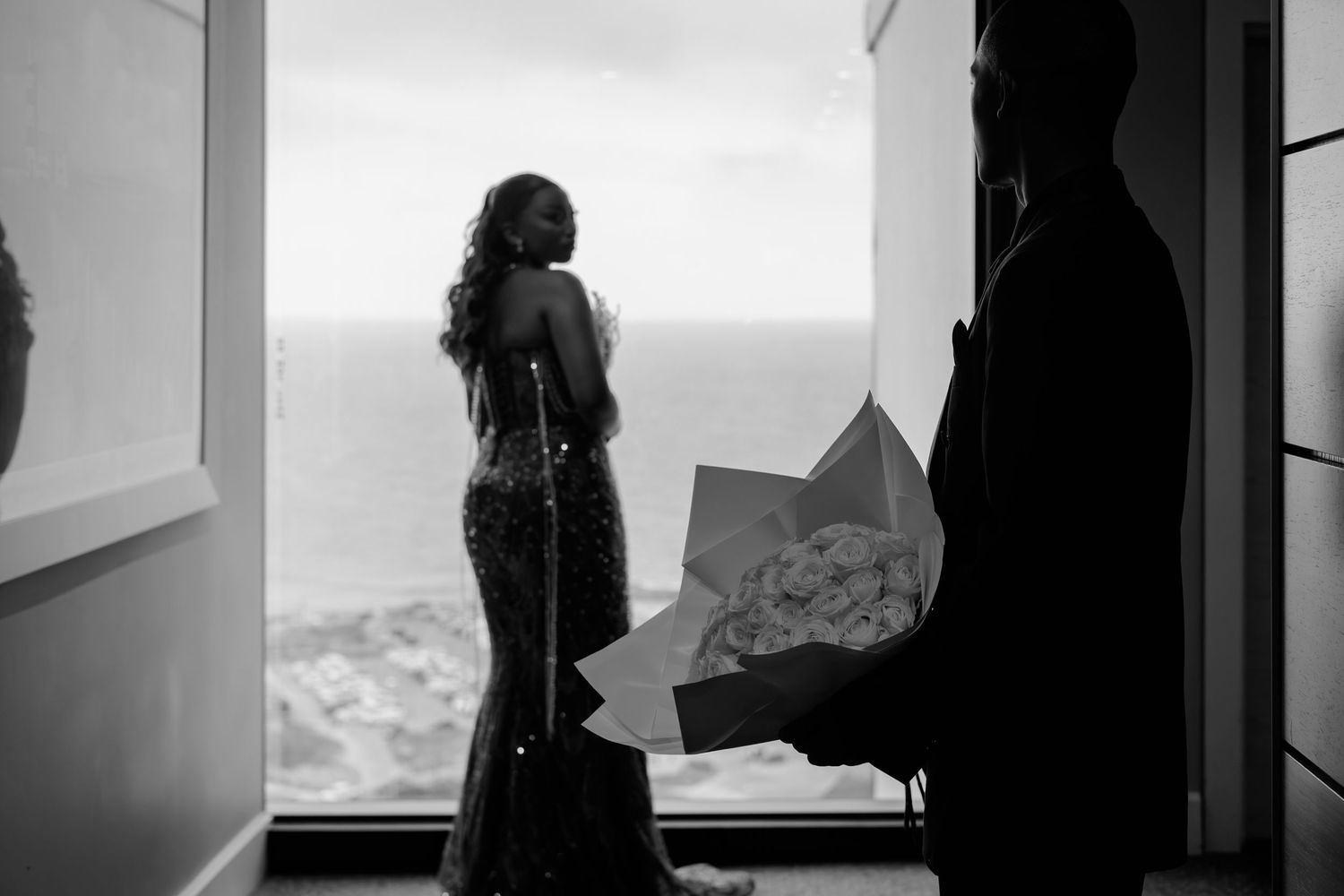 Black and white photo of a woman in an elegant gown standing by a window overlooking the ocean, man holding flowers.