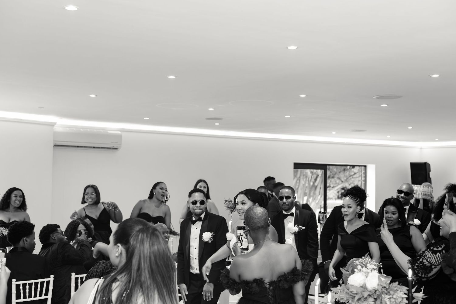 Black and white photo of lively wedding reception with guests celebrating in elegant venue.