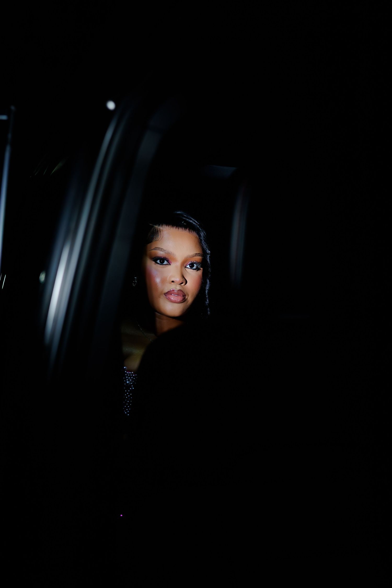A woman with dramatic makeup emerges from a dark vehicle at night, wearing a black outfit.