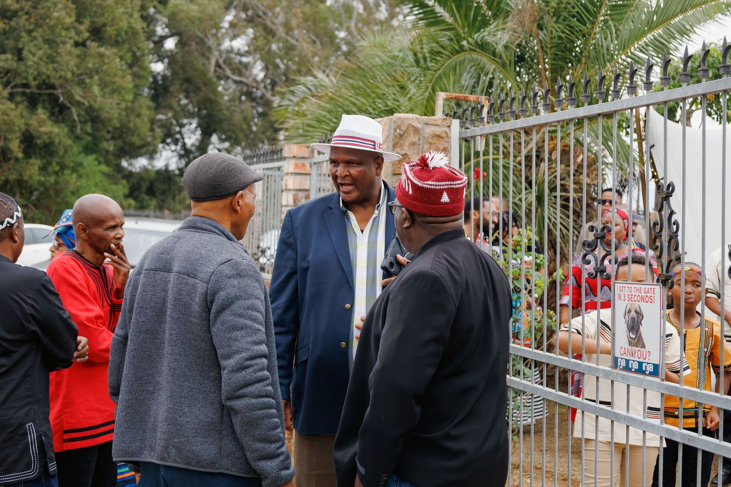 Group of men in traditional and formal attire conversing outside iron gates at a cultural gathering.