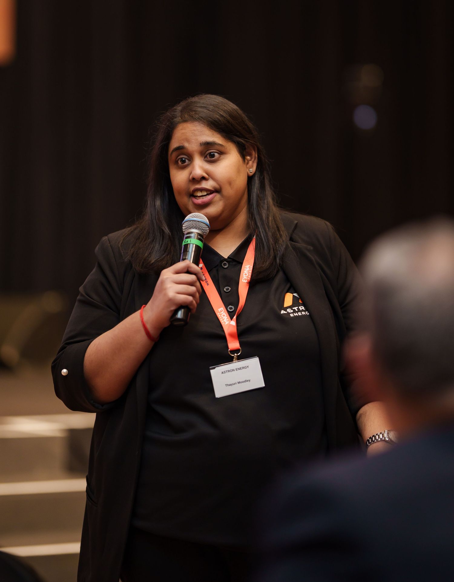 Woman in Astron Energy branded shirt speaks into microphone at retailer conference.