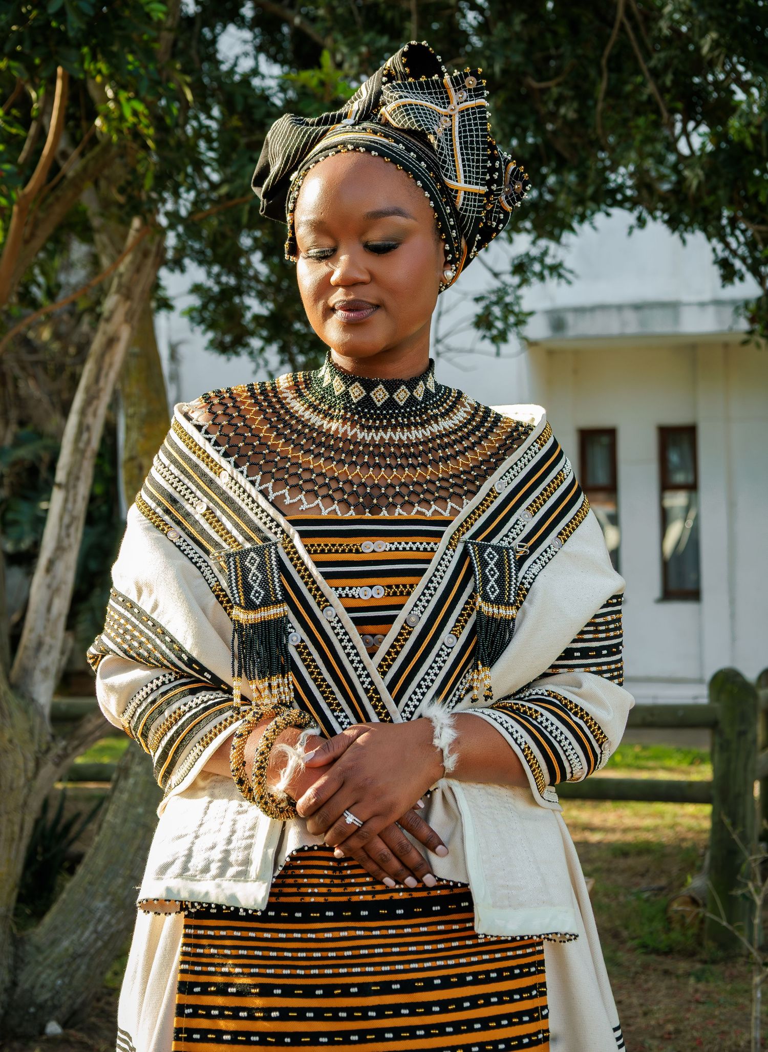 Woman in elegant traditional Xhosa dress with intricate beadwork and patterned head wrap outdoors.