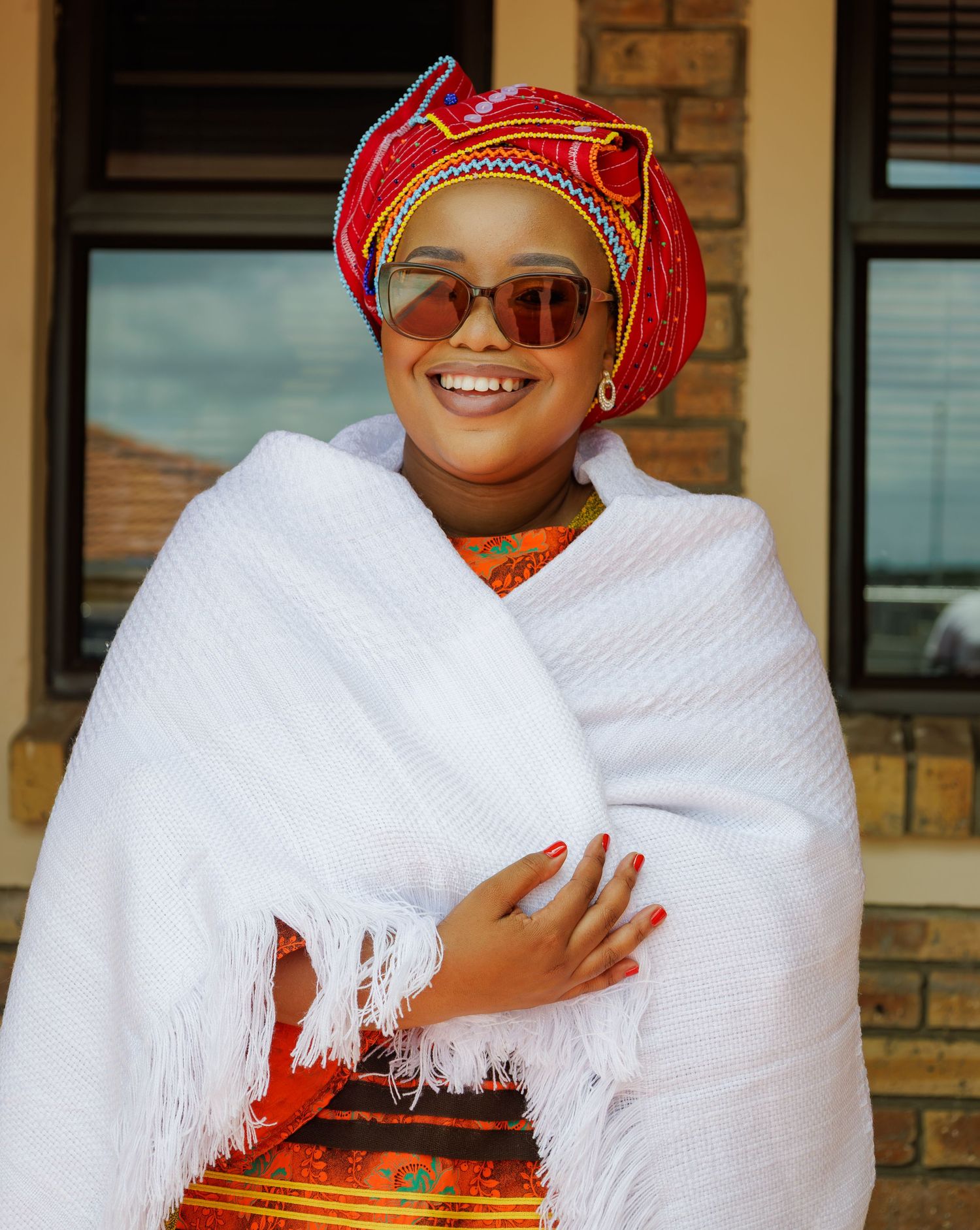Smiling woman in traditional African attire with red headwrap, white shawl, and orange dress posing outdoors.