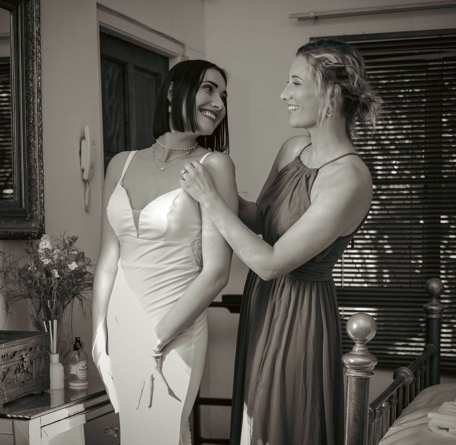 Black and white photo of bride in white gown smiling with bridesmaid adjusting her dress straps.