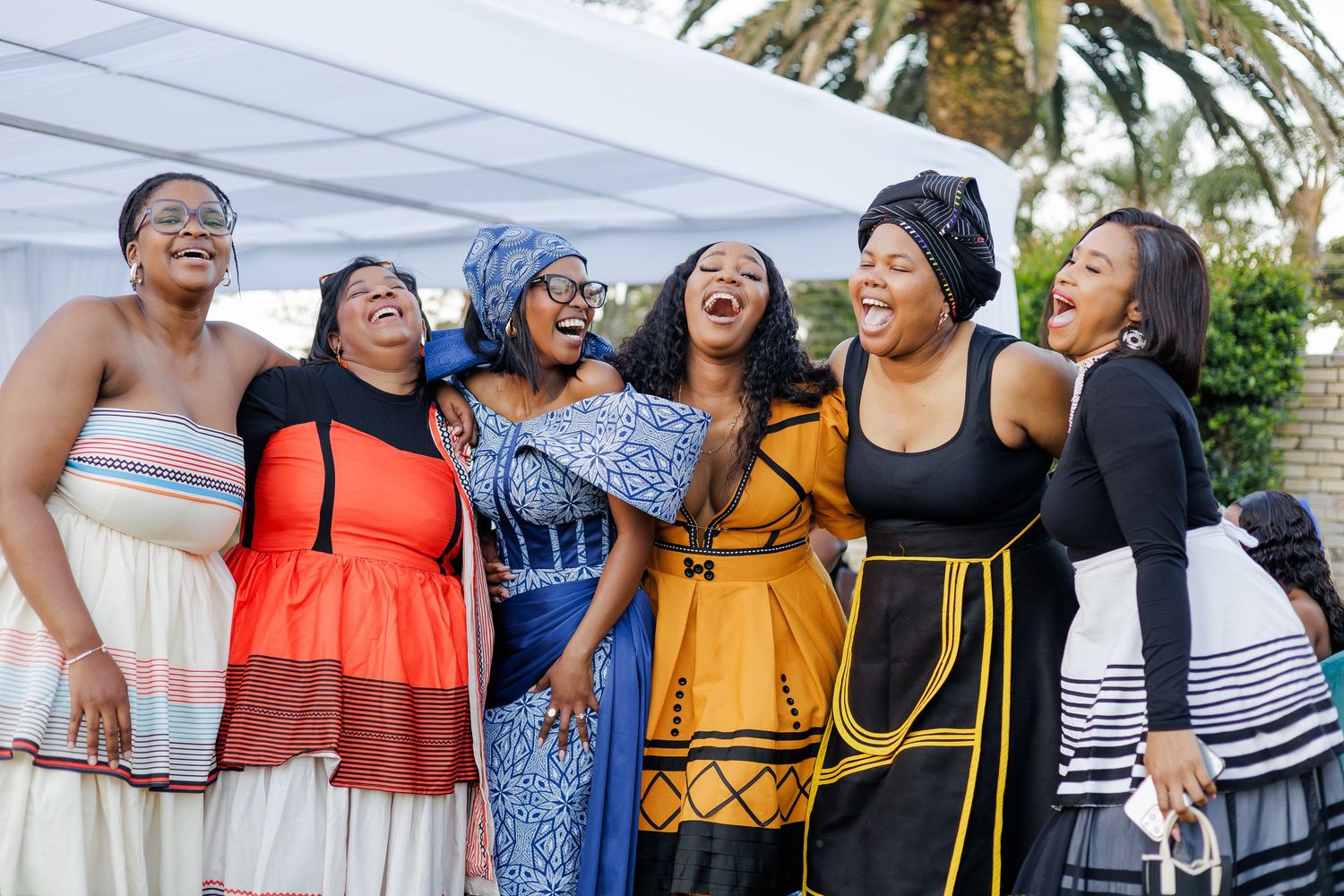 Six women laughing joyfully together at an outdoor African traditional ceremony wearing colorful cultural attire.