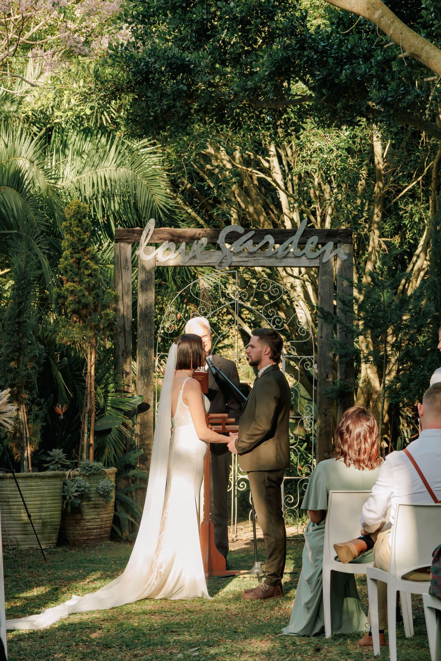 Bride and groom exchanging vows under a wooden arch in a lush garden ceremony surrounded by tropical greenery.