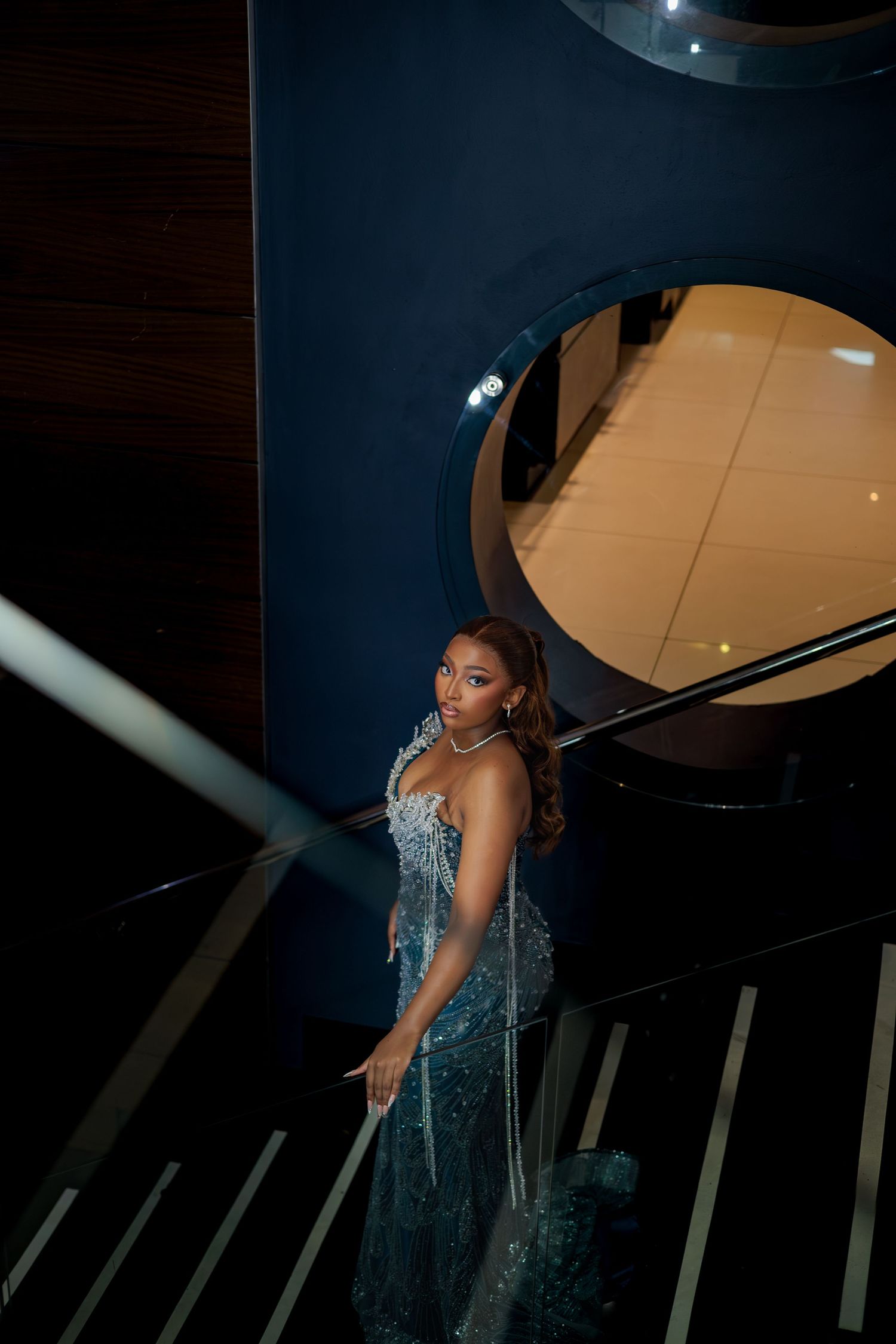 Elegant woman in a sparkling teal sequin dress smiling on a dark staircase with circular architectural detail.