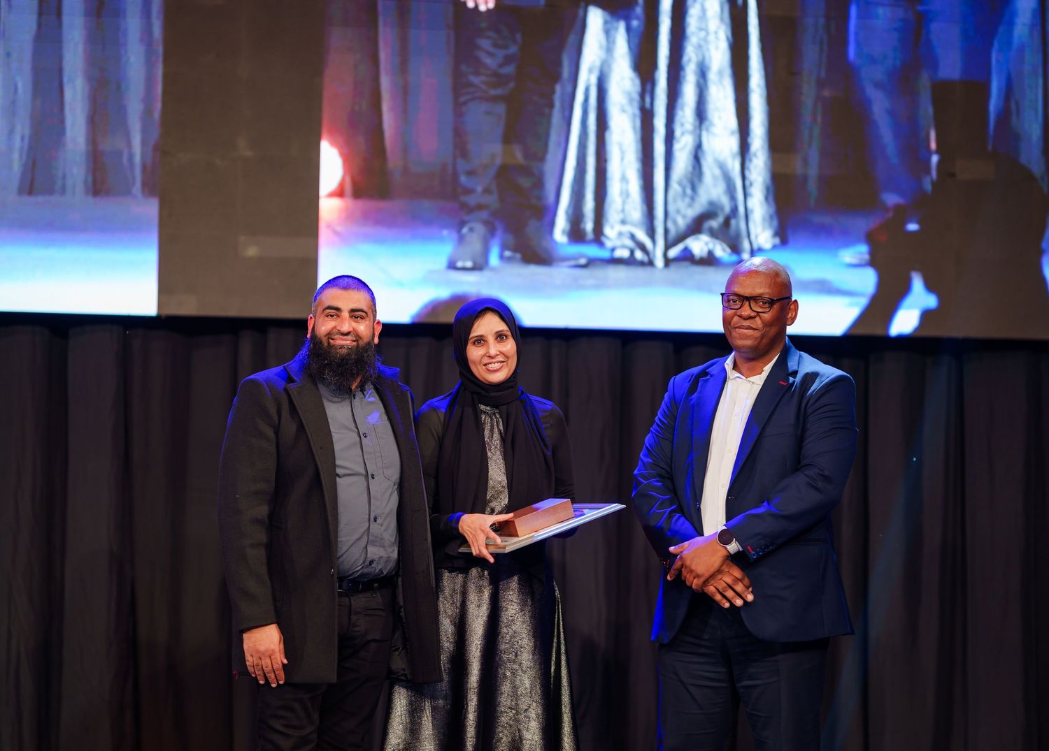 Three people pose together holding award certificate on stage at corporate gala event.
