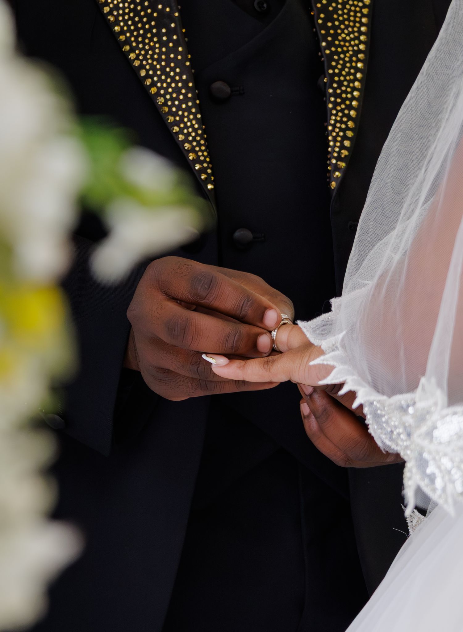 Groom placing a wedding ring on brides finger during ceremony, wearing black studded suit with white floral bouquet nearby.