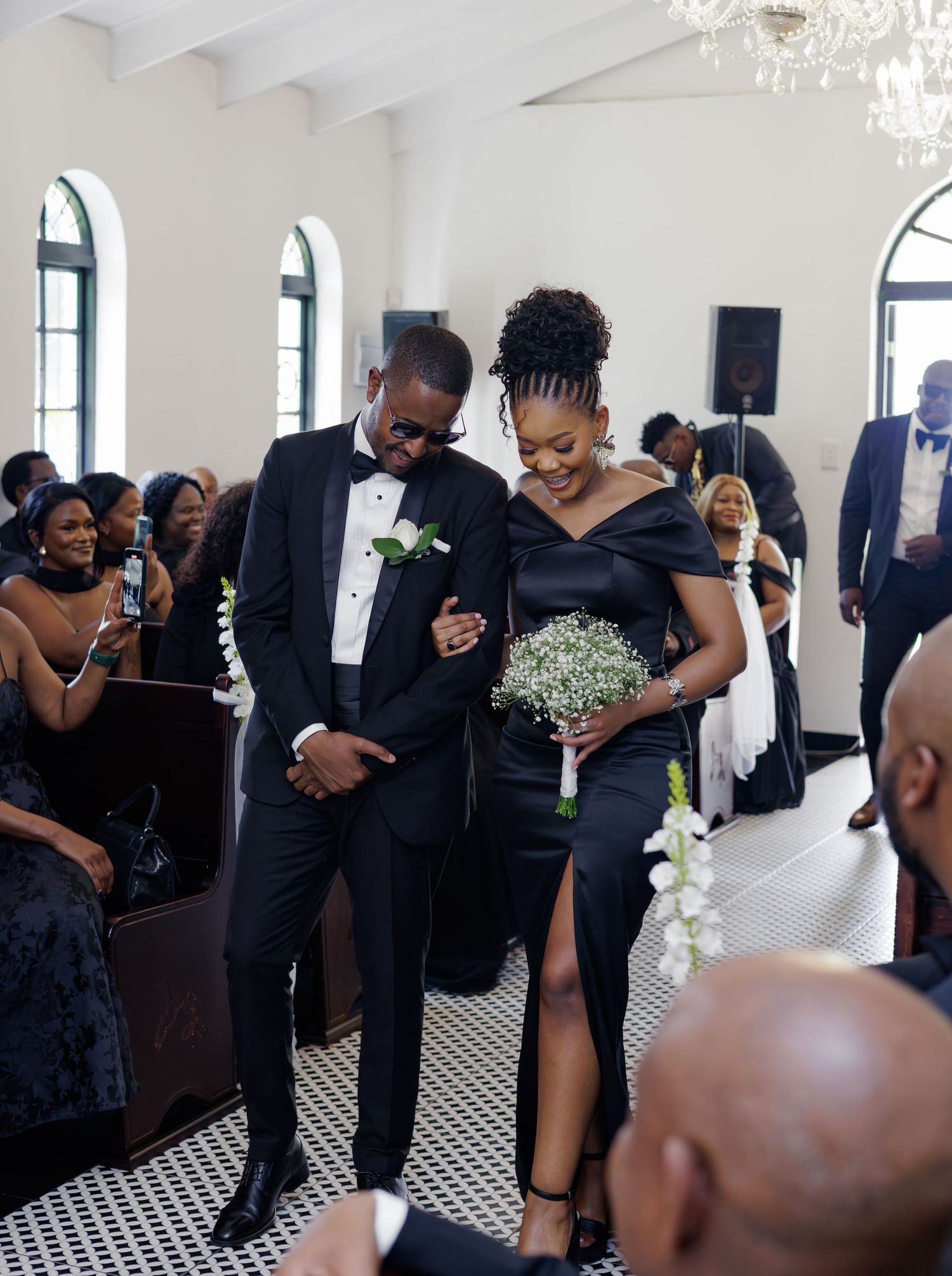 Bridesmaid and groomsman walking down church aisle in matching black wedding attire with white bouquet.