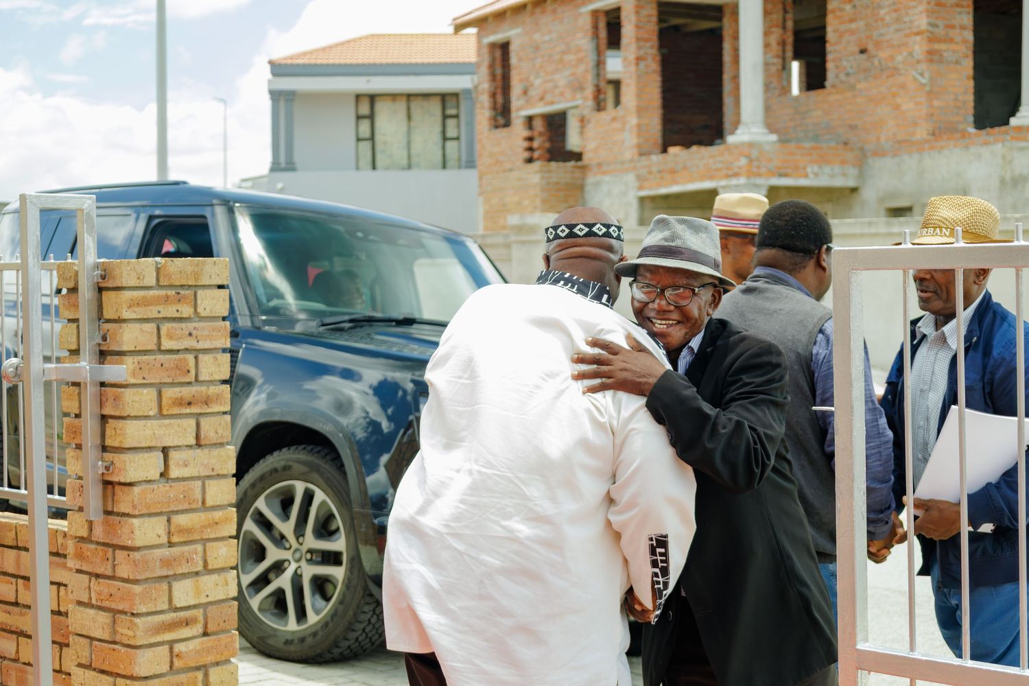 Two men sharing a warm embrace near a blue SUV outside a brick building during a gathering.