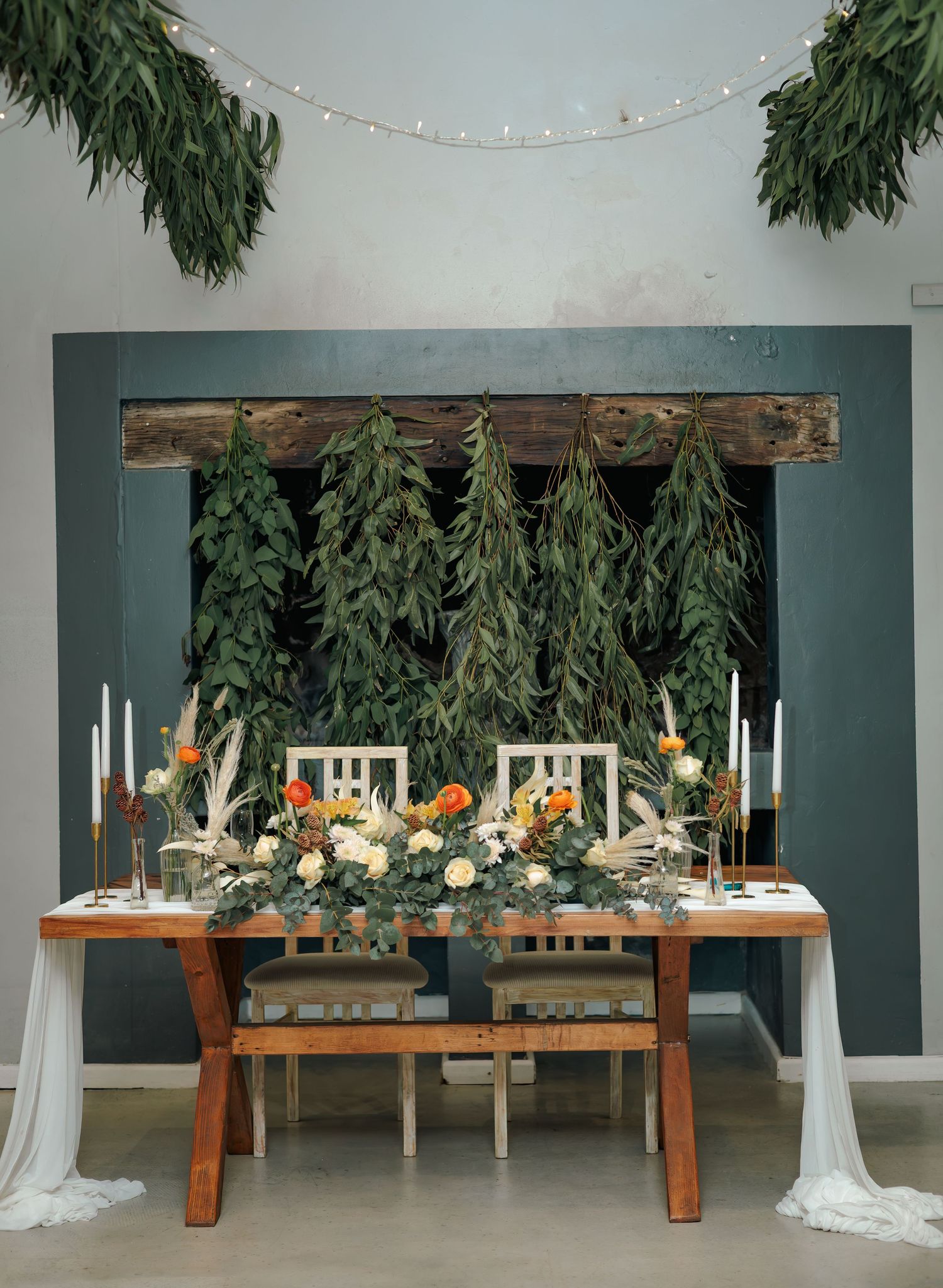 Rustic wedding sweetheart table with floral centerpiece, candles, and hanging greenery backdrop decor.