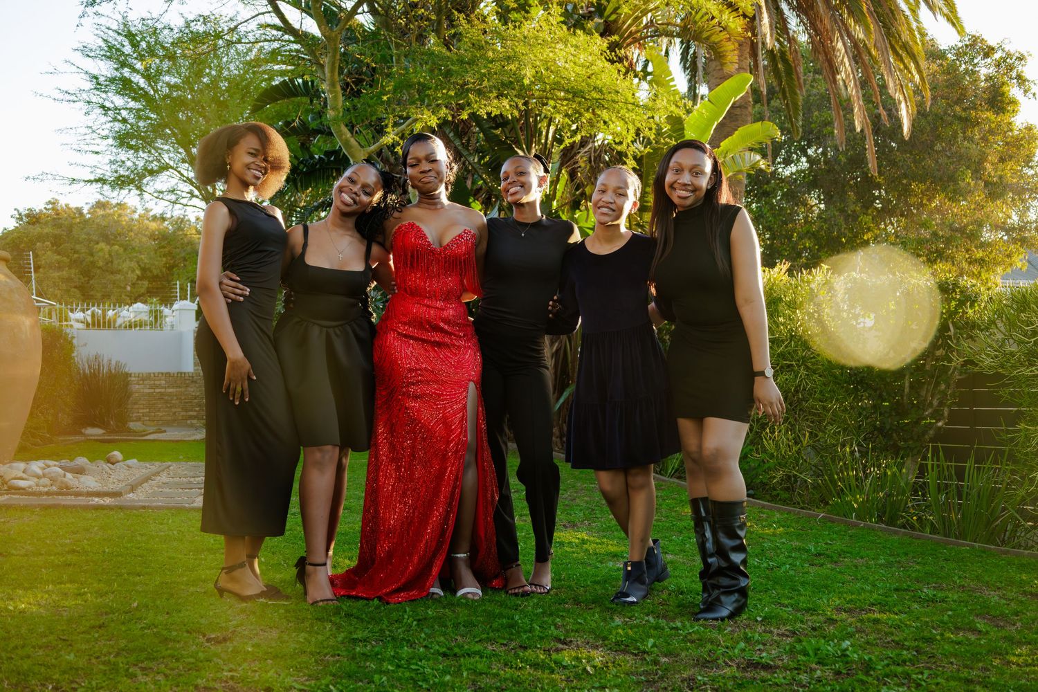 Six young women in formal attire pose together outdoors, one in a red gown surrounded by friends in black dresses.