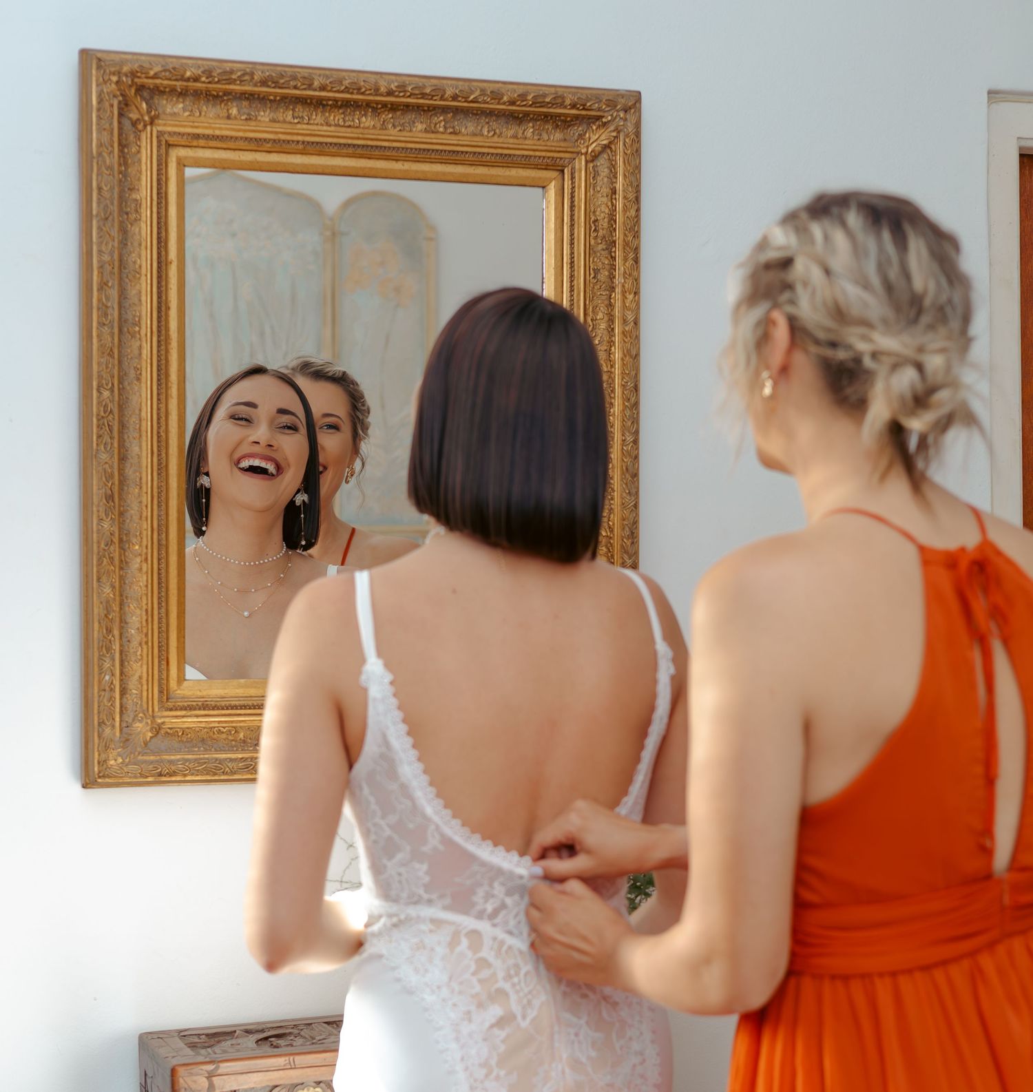 Bride in white lace gown laughing at mirror reflection while bridesmaid in orange dress fastens her dress.