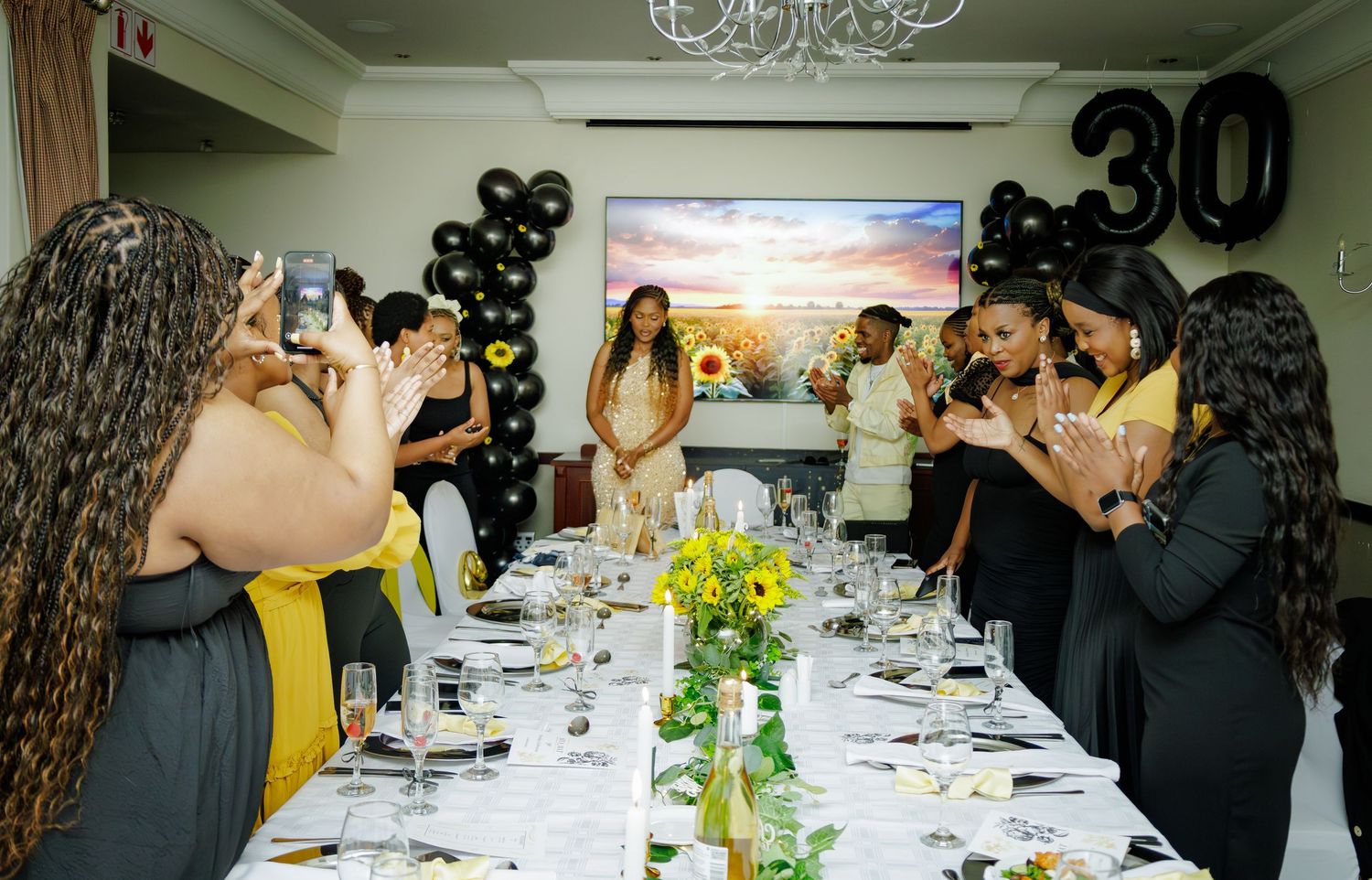 Guests applauding at a 30th birthday dinner party with black balloons and sunflower themed decorations.