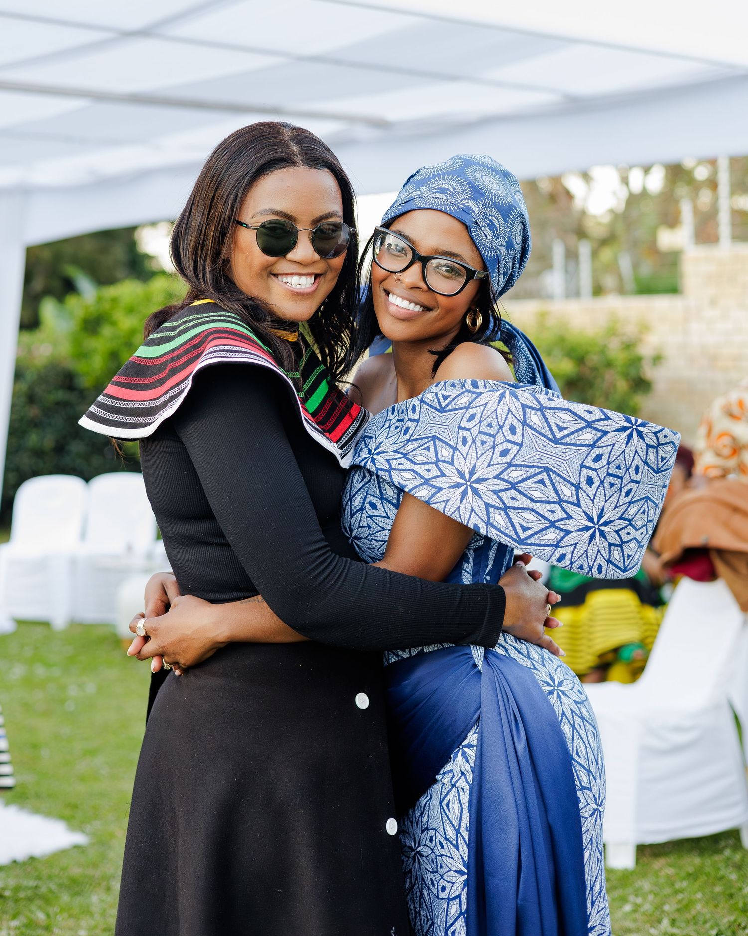 Two women smiling and embracing at outdoor African celebration wearing traditional Xhosa and Ndebele attire.
