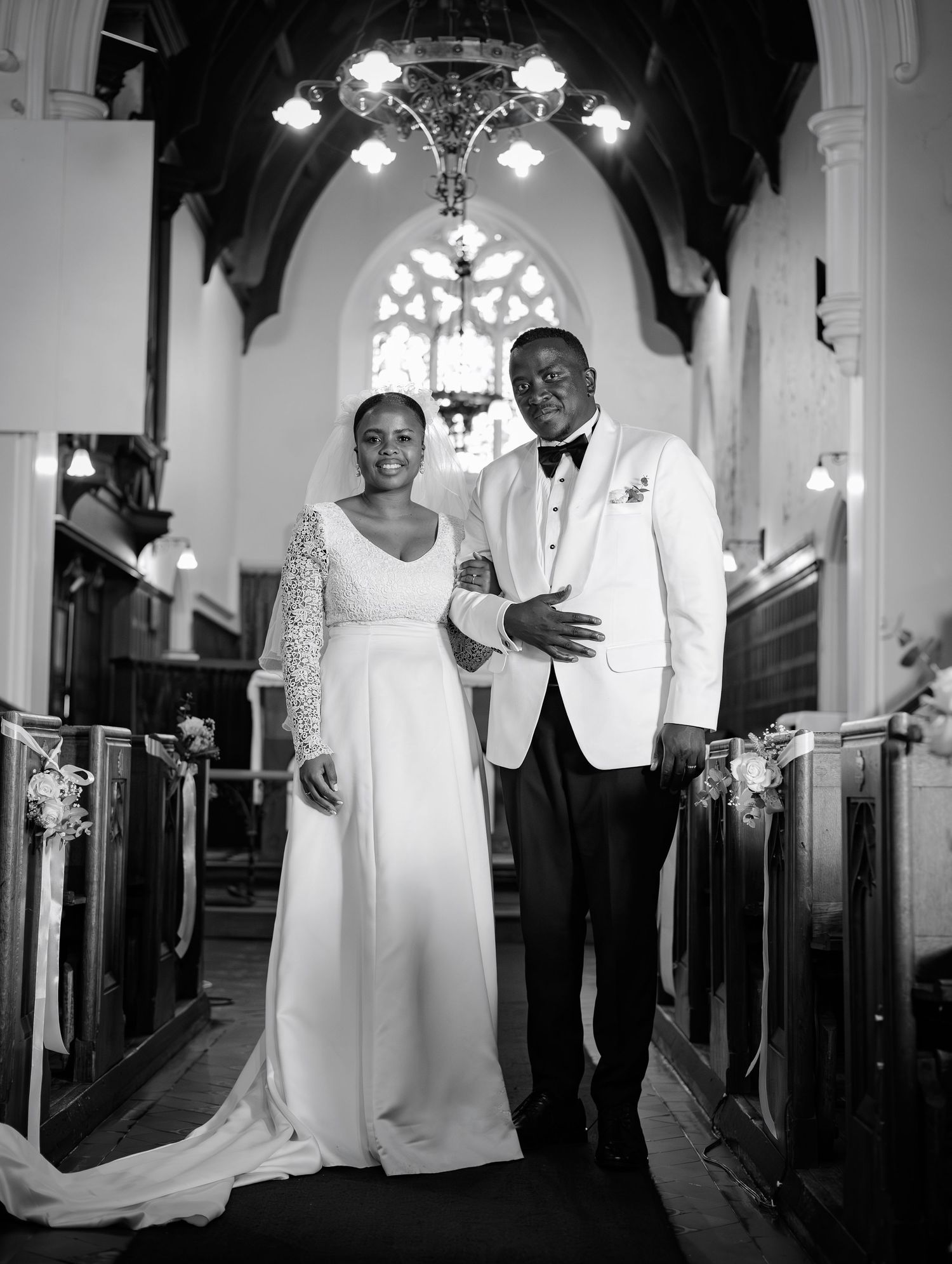 Black and white portrait of a smiling bride and groom posing together in a beautifully decorated church aisle.