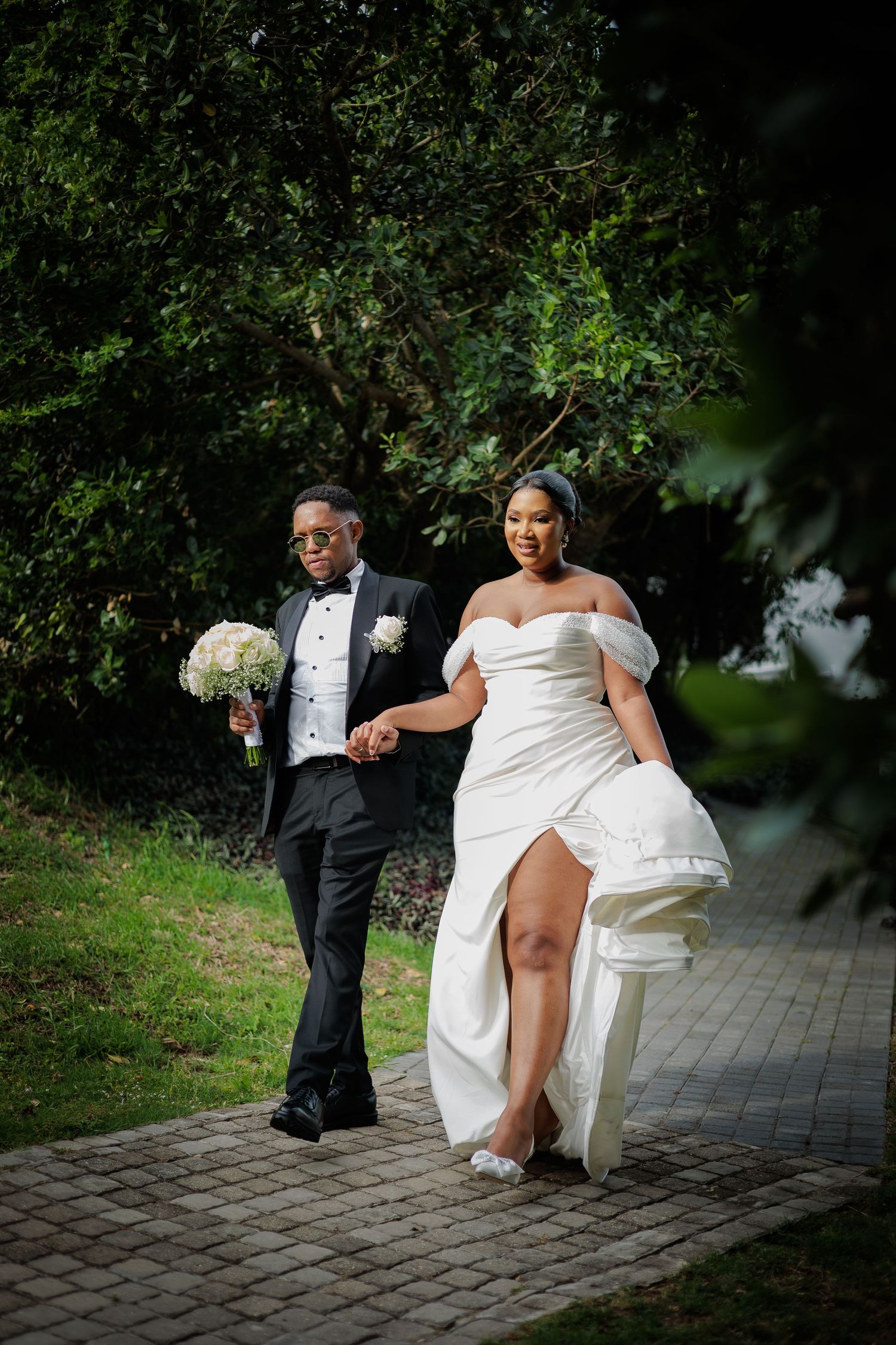 Bride in white slit gown and groom in tuxedo walk together through garden pathway.