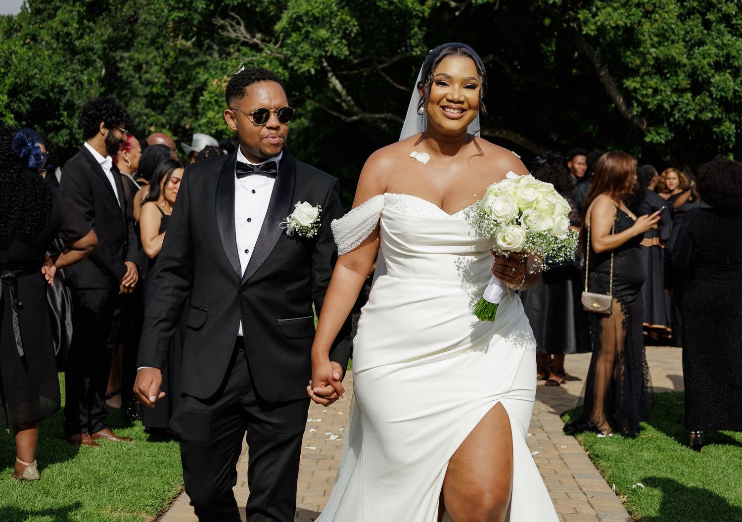 Newlyweds walk hand-in-hand outdoors, bride in white off-shoulder gown holding bouquet.