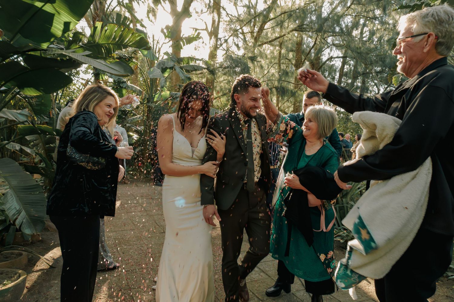 Newlyweds celebrate with confetti toss as smiling wedding guests shower them with colorful paper outdoors.