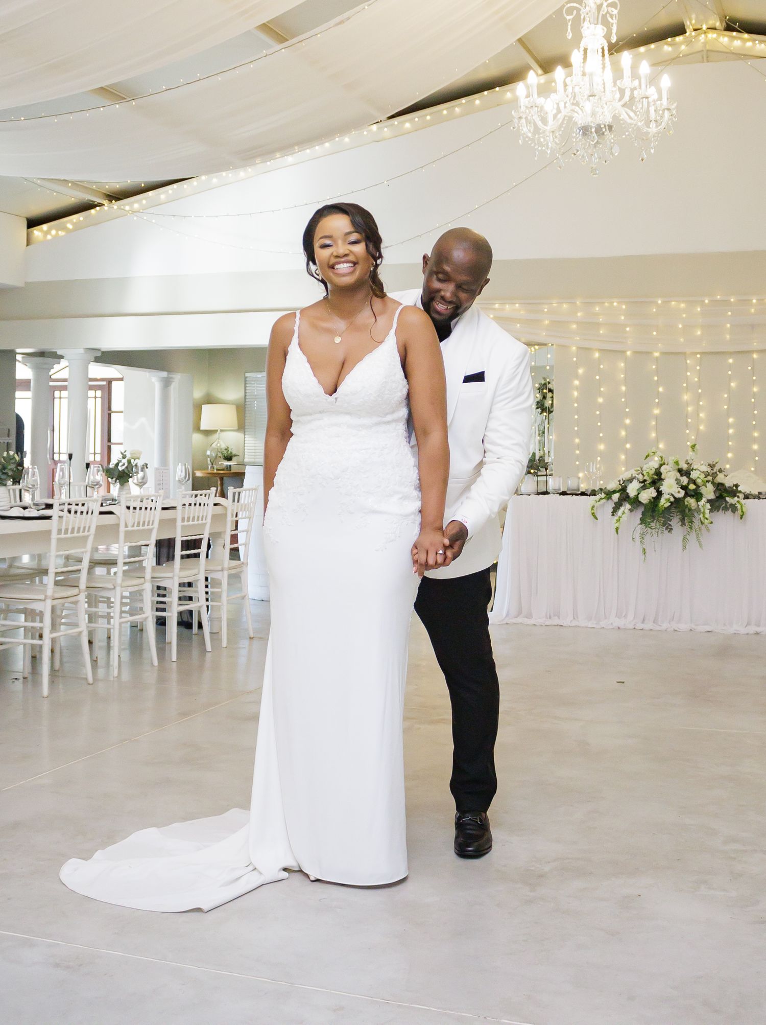 Happy newlyweds holding hands and smiling in a fairy-light decorated wedding reception hall.