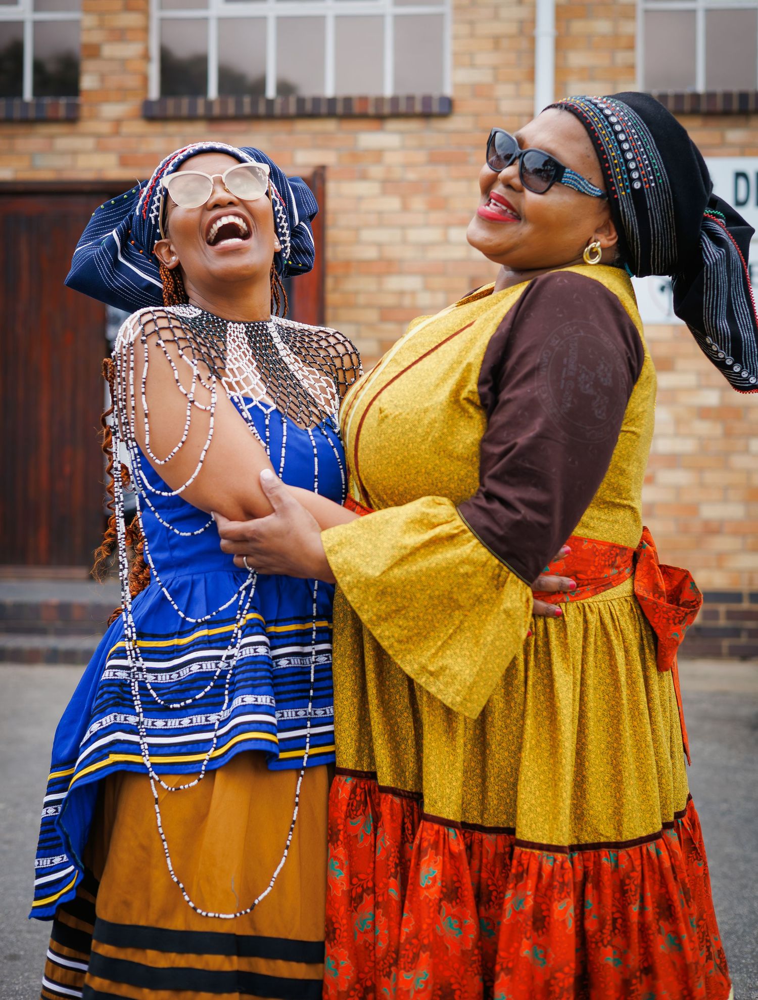 Two women laugh joyfully while wearing vibrant traditional African dresses with beaded accessories outdoors.