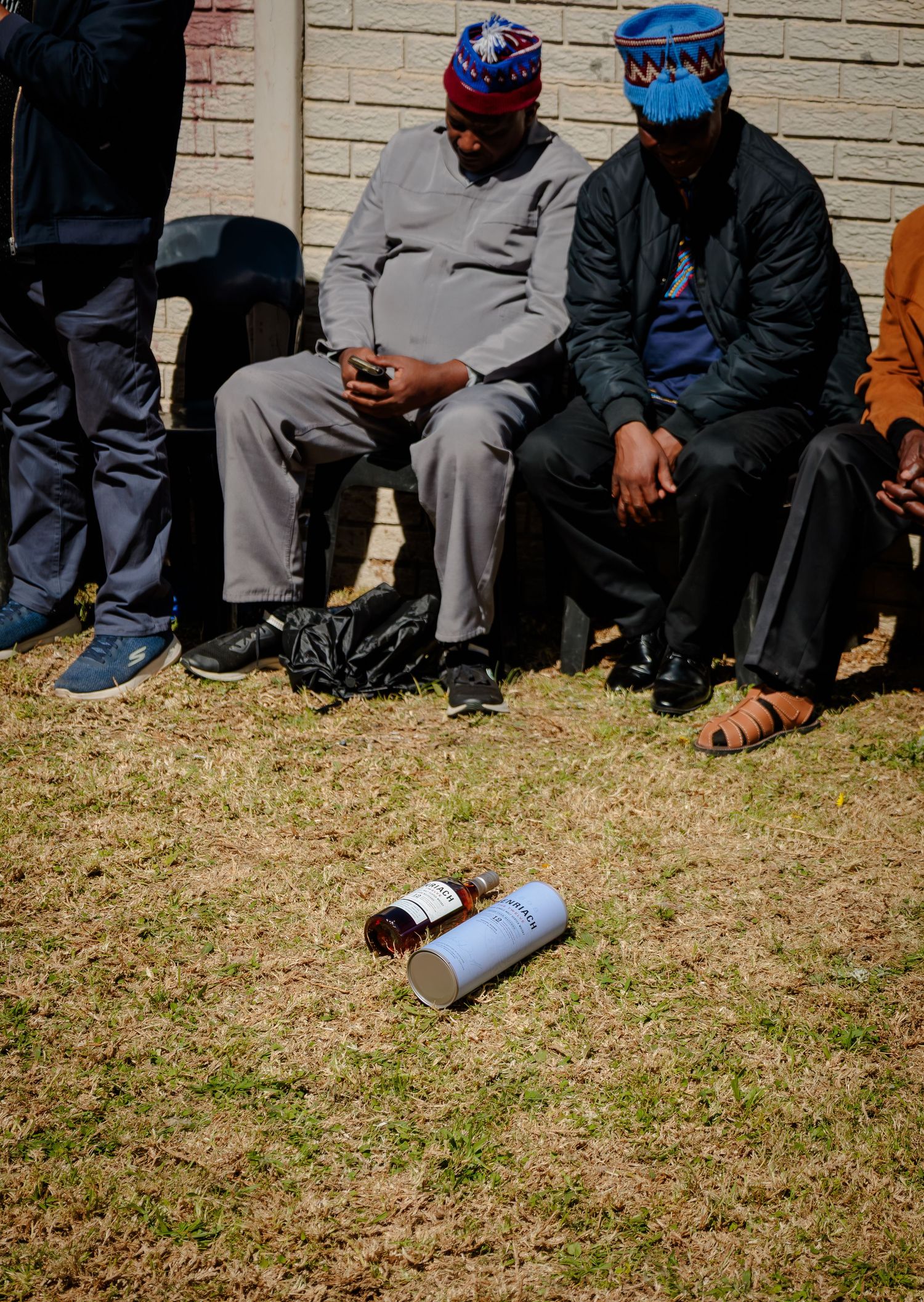 Two men wearing traditional Xhosa beaded caps sit outdoors with a bottle on the grass before them.