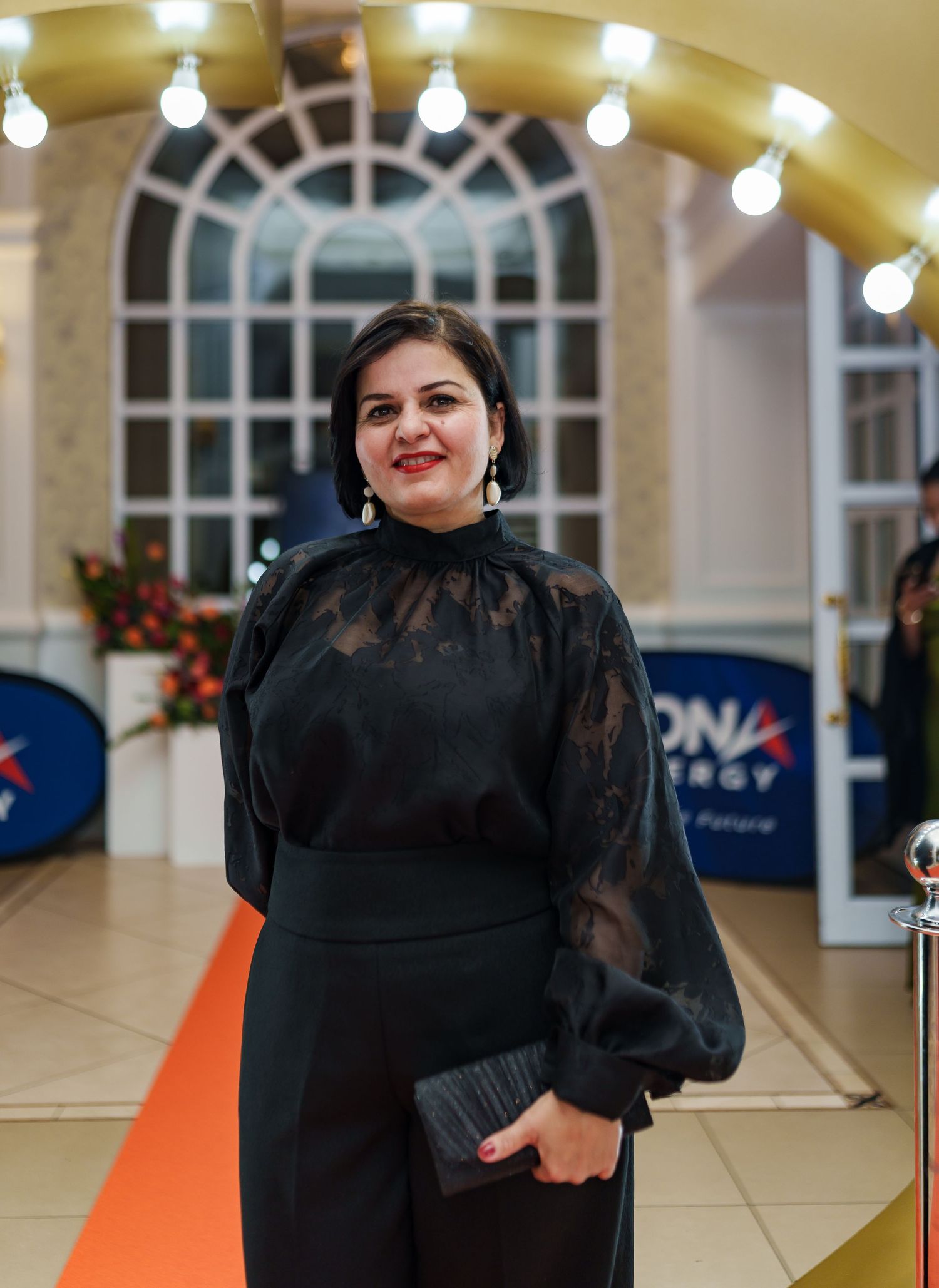 Elegant woman in black lace top holding clutch on orange carpet at corporate gala.