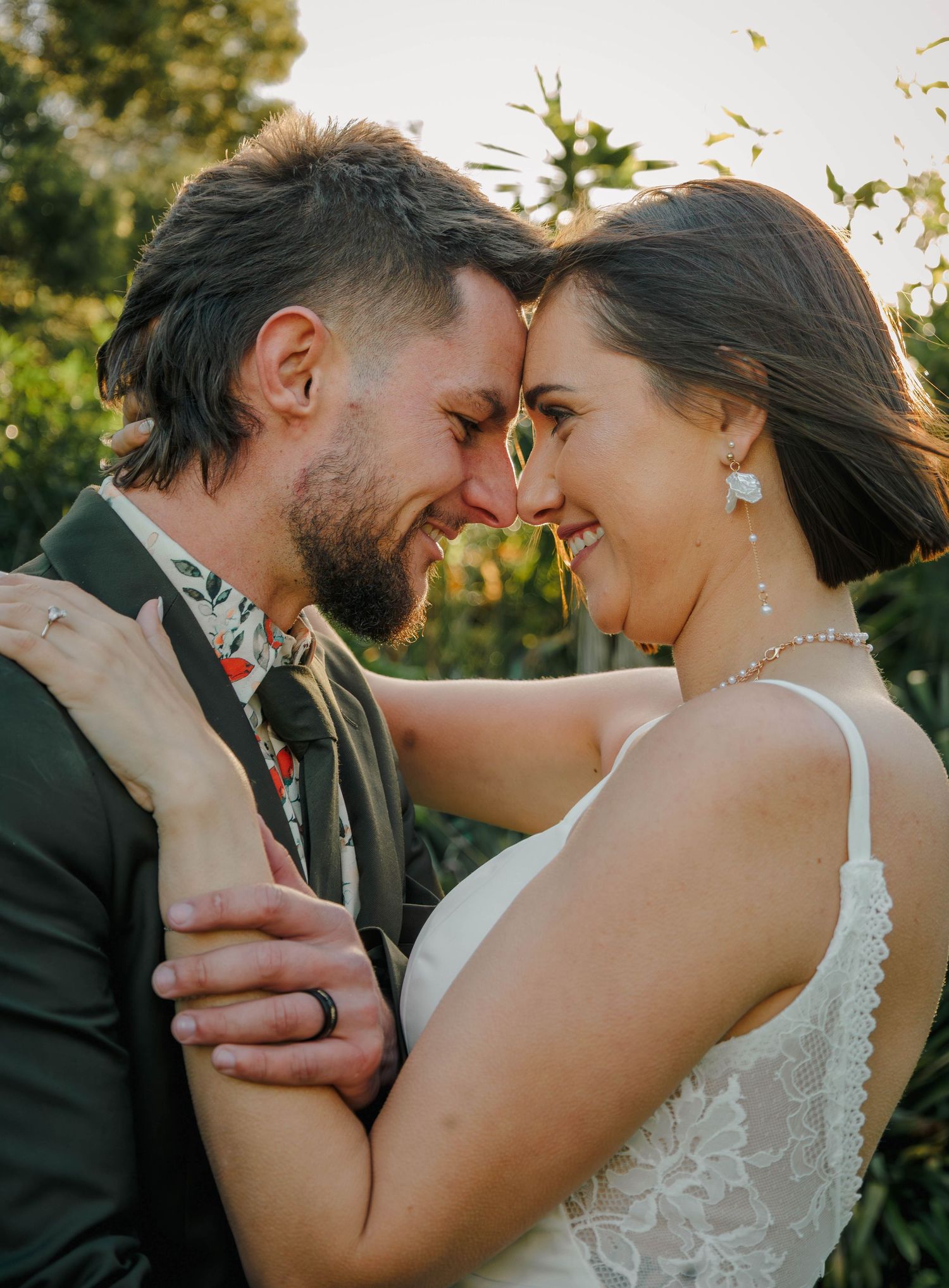 Bride and groom touch foreheads smiling intimately in golden sunset light among garden greenery.