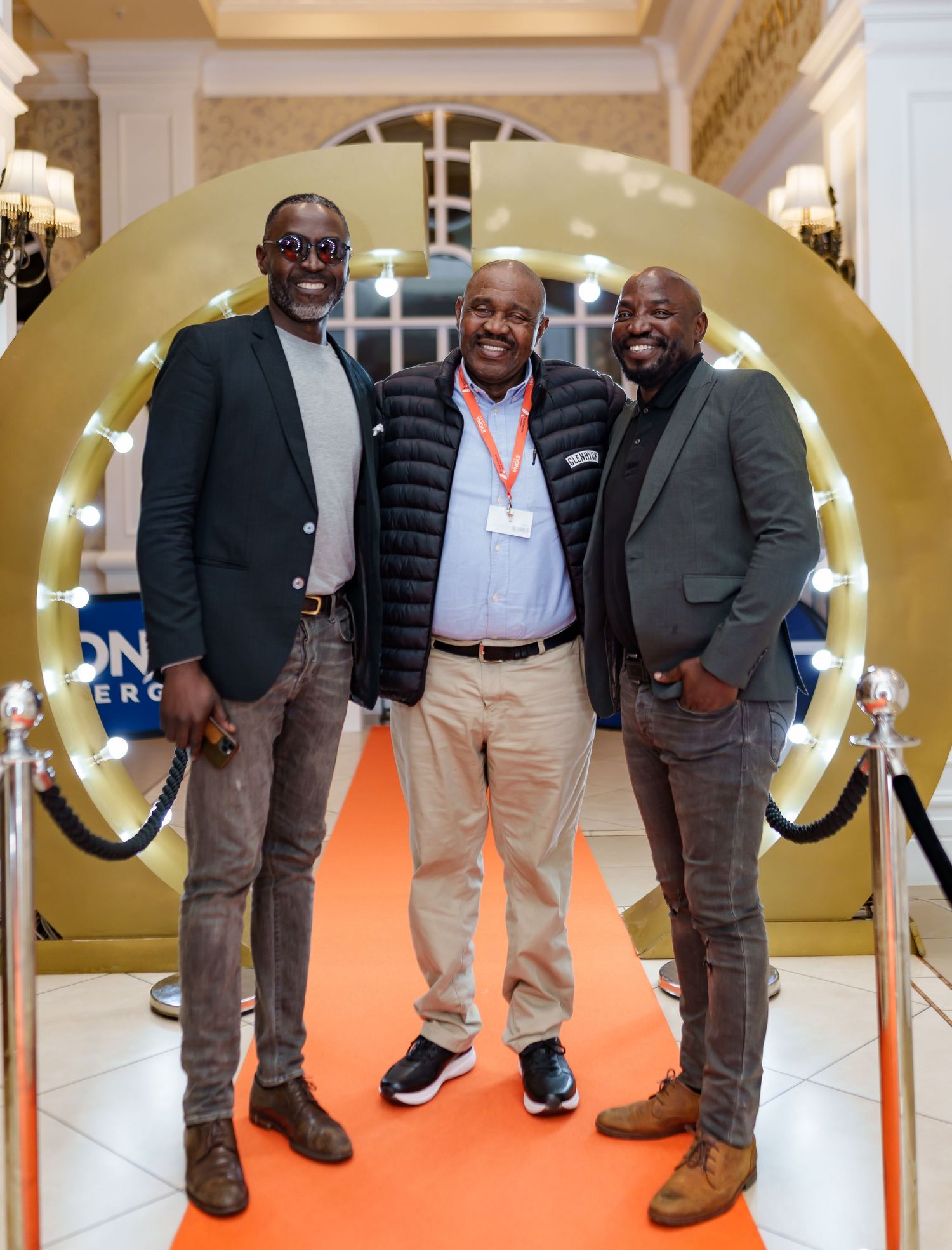 Three men smiling together on orange carpet at Beyond Energy event entrance.