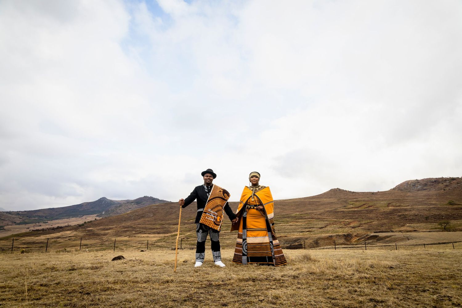 Couple in traditional Basotho blankets holding hands in a vast mountain landscape under cloudy skies.