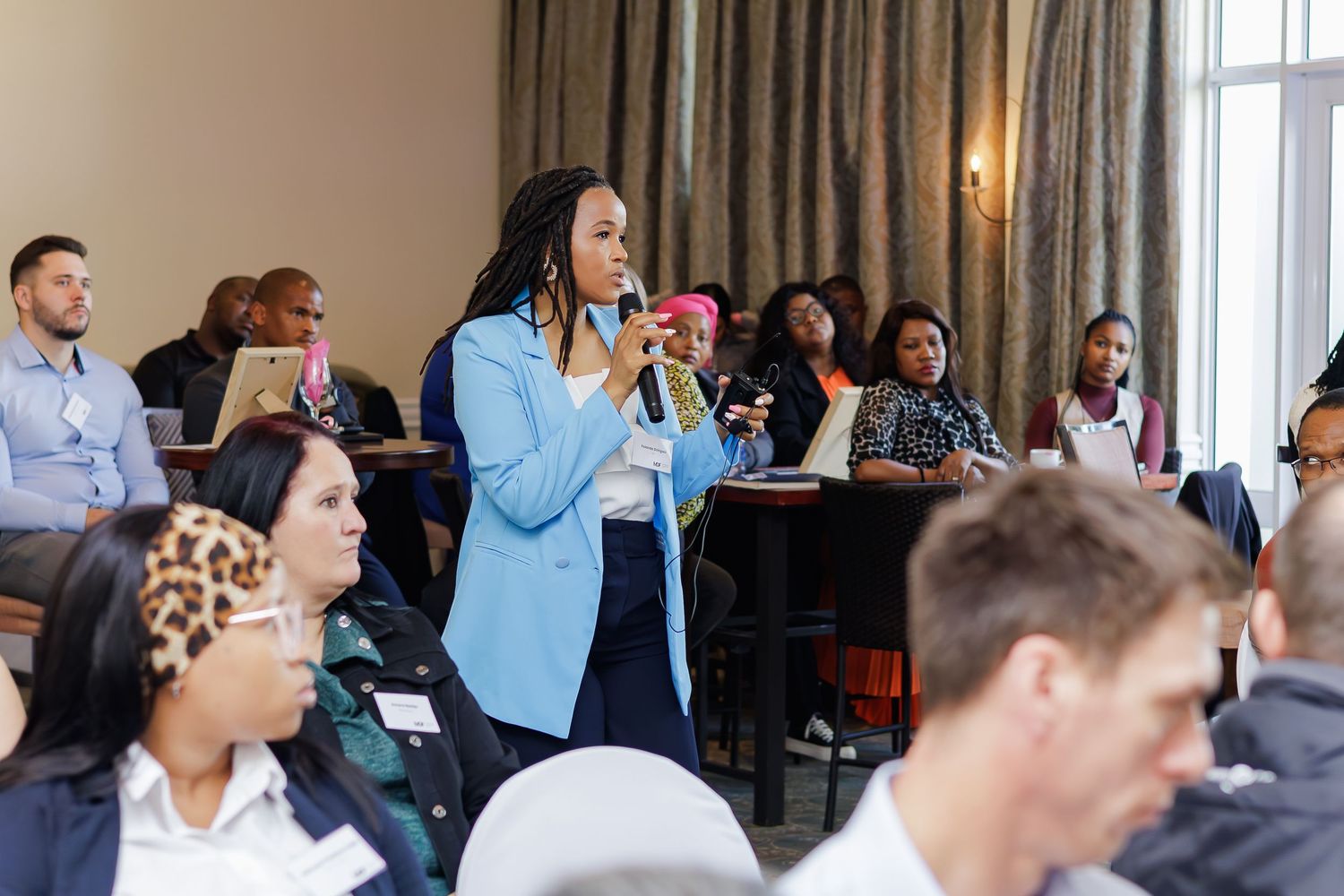 Woman in blue blazer speaking into microphone at a diverse professional conference or seminar.