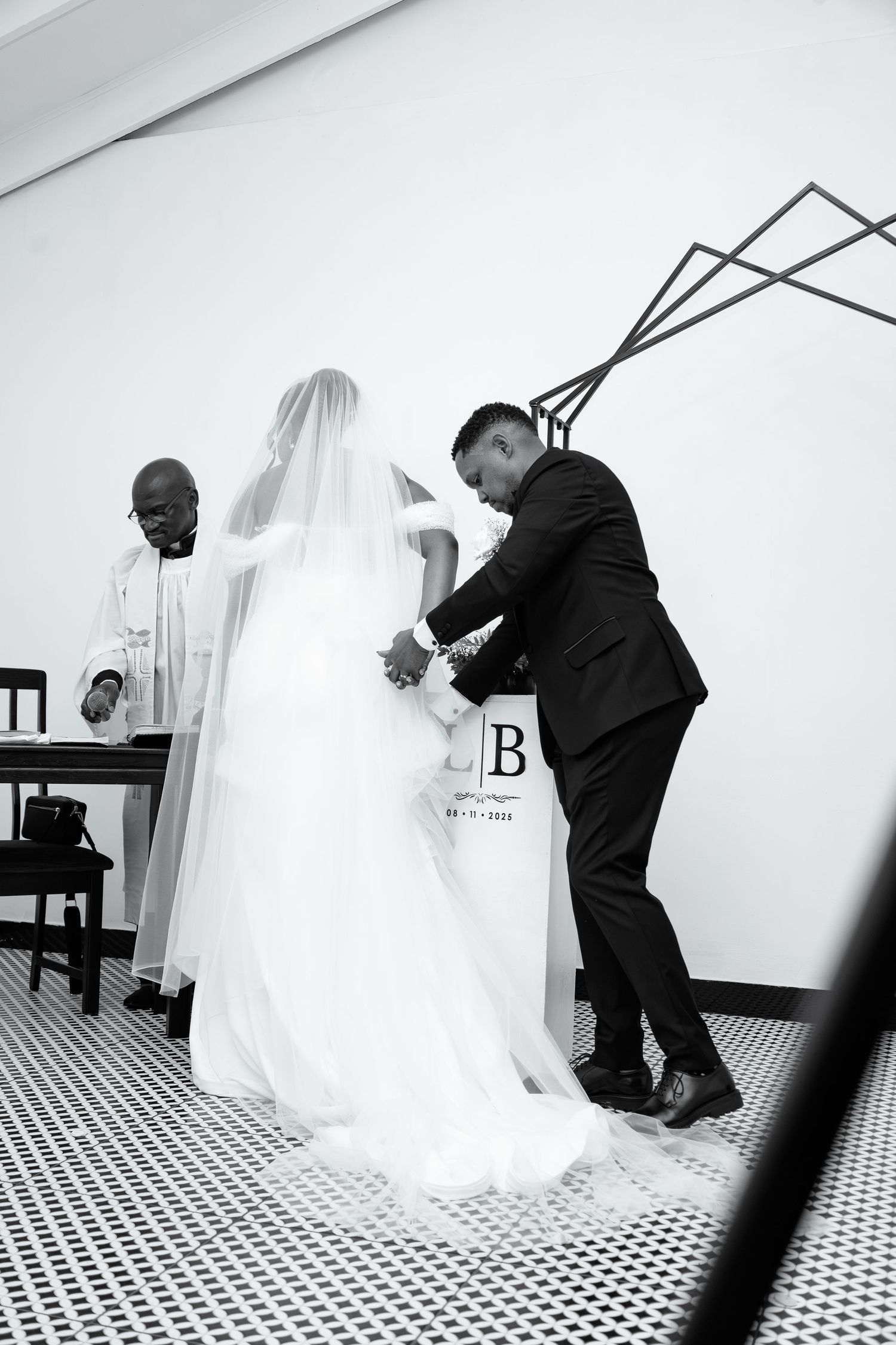Black and white photo of groom adjusting bride's wedding dress train as officiant watches.