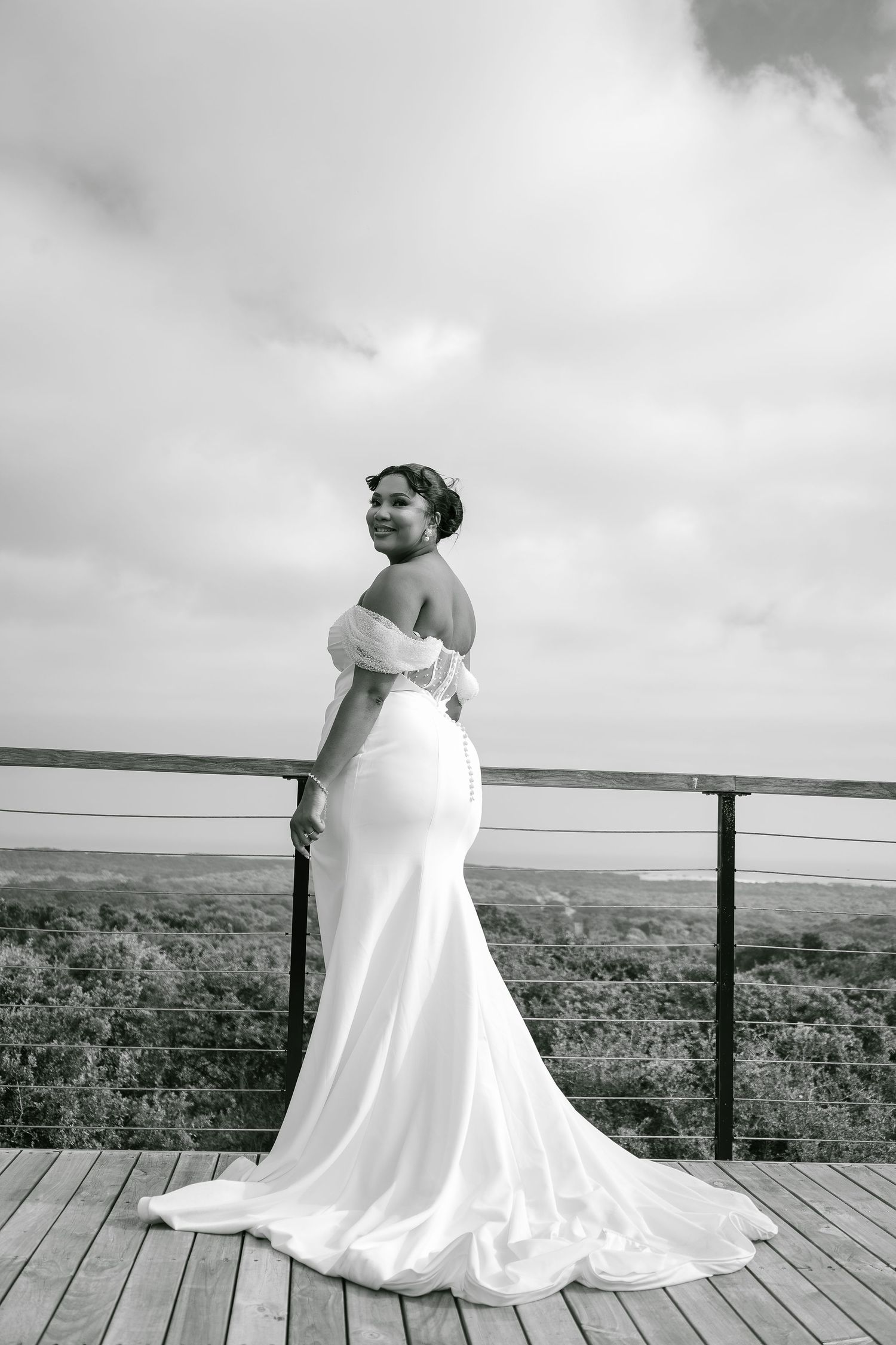 Black and white portrait of a bride in an off-shoulder white gown on an outdoor deck.